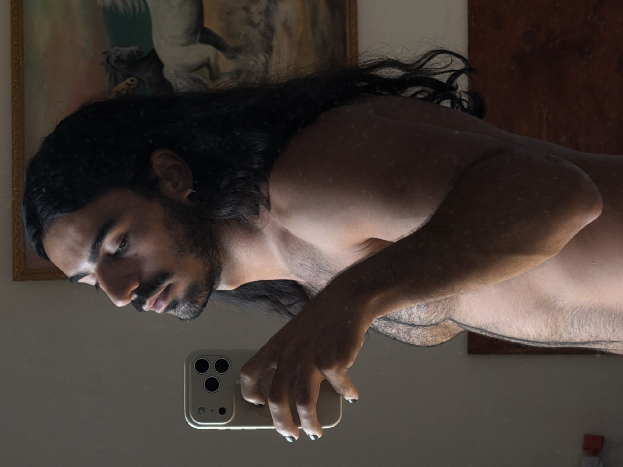A person with long dark hair and a mustache is taking a mirror selfie in a room. They are holding a silver smartphone in their right hand and looking at the reflection. The person is shirtless and their skin has some light spots. In the background, there is a painting of horses on the wall and a wooden headboard.