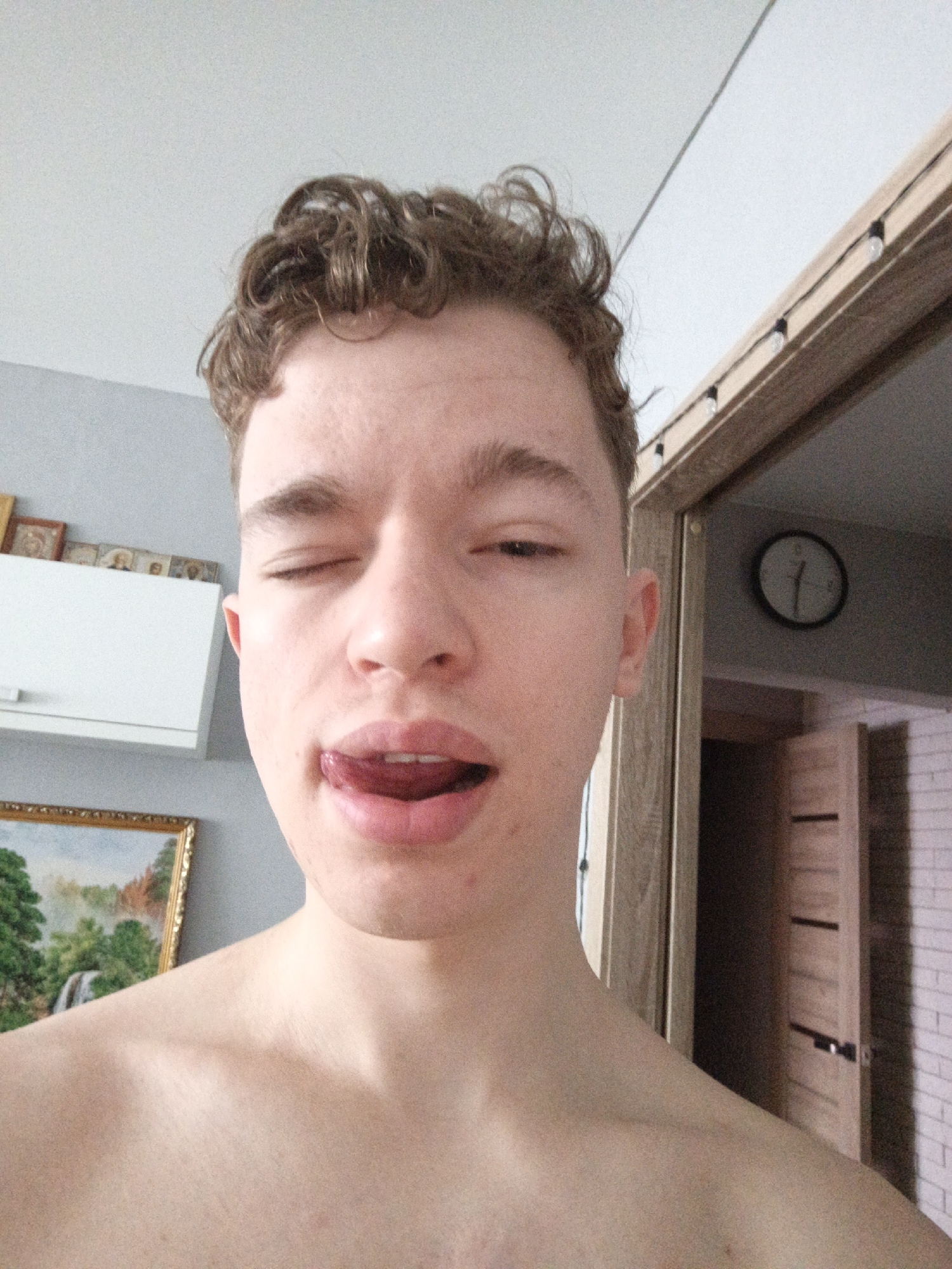 A shirtless young man with curly brown hair is posing for a selfie indoors. He is partially winking with his tongue sticking out. Behind him is an open doorway, revealing a clock on the wall and a glimpse into another room with a white brick wall and a wooden door. To the left, a white cabinet hangs above a framed picture. It is impossible to identify the city of Chaikovsky, Russia as the location, since there are no geographical landmarks.