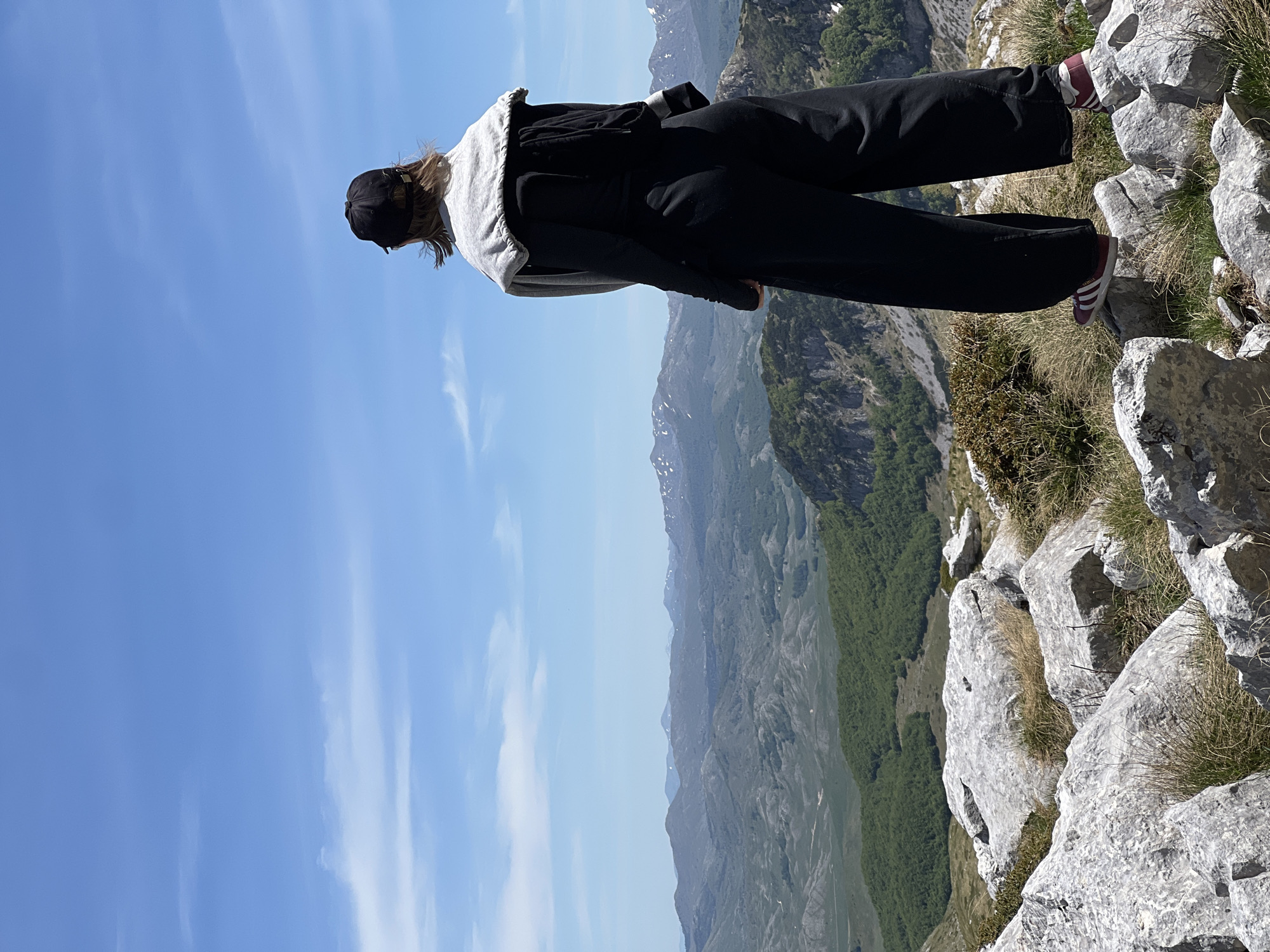 A person is standing on a rocky outcrop in Pašina voda, Montenegro, with a vast mountainous landscape visible in the background. The person is wearing a black cap, a gray and white hoodie, black pants, and maroon sneakers. They are positioned near the edge of the rocky terrain, which includes grass and other vegetation. The background displays a series of mountain ranges under a blue sky with streaky clouds.