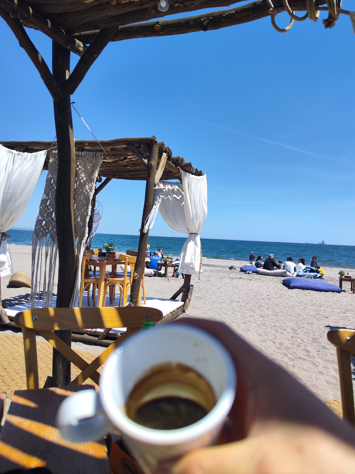 On a sunny day in Burgas, Bulgaria, a person holds a cup of coffee in the foreground, blurring the details of the beverage. In the midground, a beachside setting is arranged with a wooden gazebo draped with white fabric and macrame decorations, under which a wooden table and chairs are placed. Further along the beach, people are seated on large, blue cushions. The sea stretches out to the horizon under a clear blue sky.