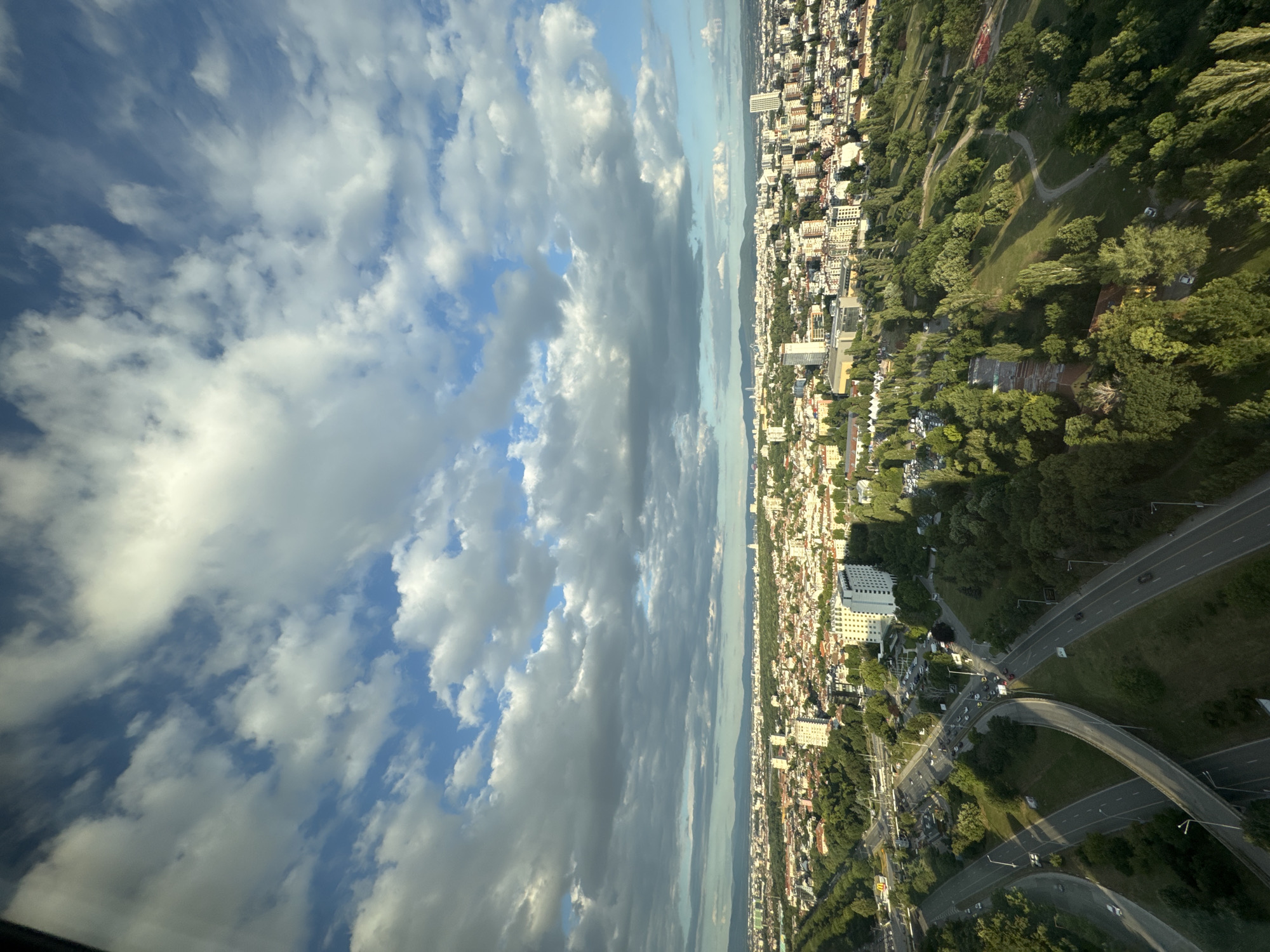 This is an aerial view of Sofia, Bulgaria, looking northeast. The foreground features a complex highway interchange with several lanes of traffic moving in various directions. The highway is surrounded by lush green trees and grass.
In the mid-ground, the city of Sofia sprawls out, characterized by a dense collection of residential and commercial buildings with light-colored facades. Several taller, modern buildings, including at least one prominent white high-rise hotel, punctuate the cityscape. The city is interspersed with numerous green spaces and parks, indicating a well-planned urban landscape with a significant amount of vegetation.
The background shows the distant outlines of hills or mountains, softened by atmospheric perspective. The sky is a dominant feature, filled with a dramatic display of white and gray cumulus clouds against a bright blue sky, suggesting dynamic weather conditions. The lighting indicates it is likely late afternoon or early evening, with sunlight casting shadows and highlighting the textures of the clouds and the urban environment. There are no visible people in the image.