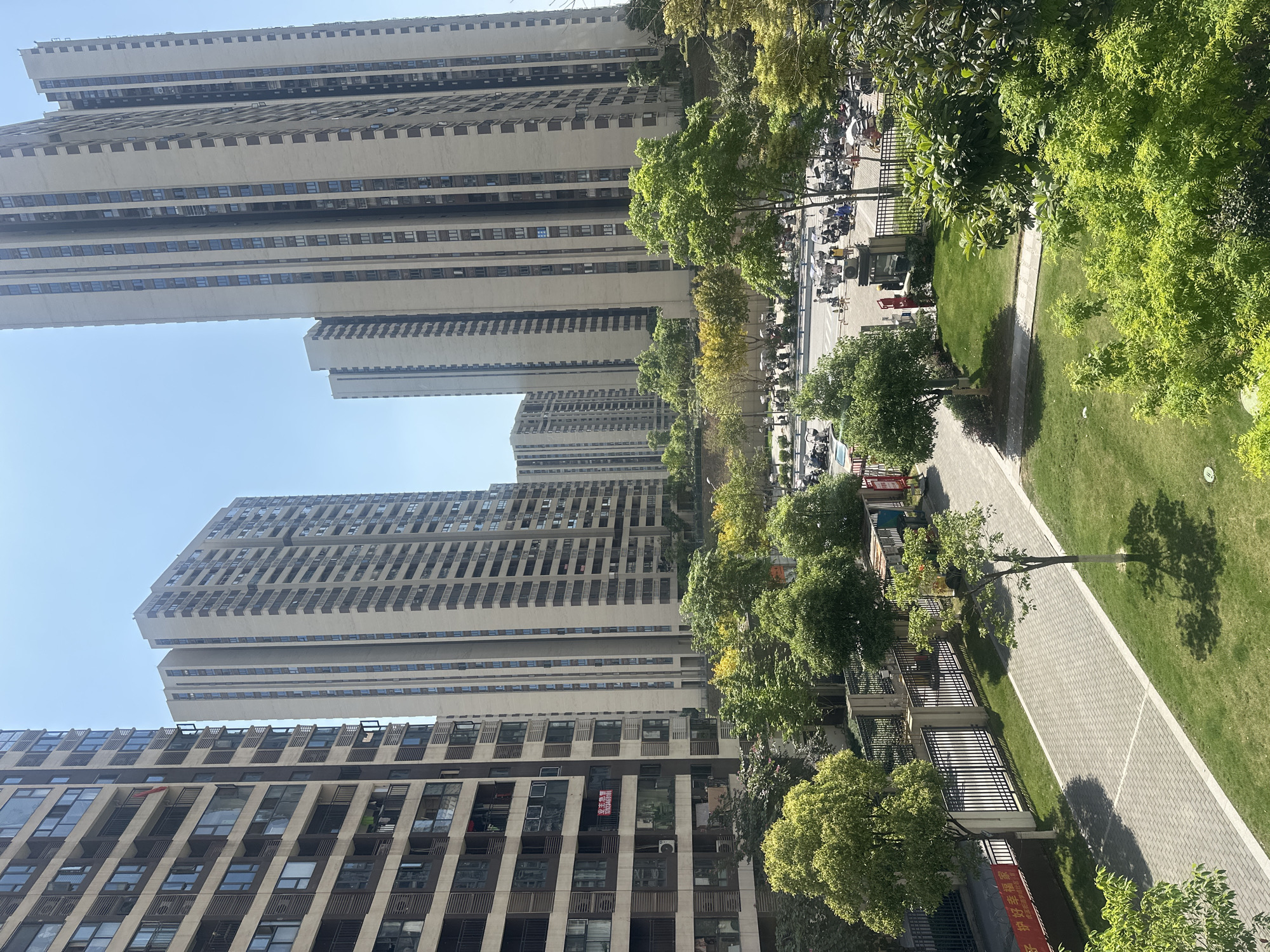 The image shows a densely populated urban area, likely in the Chongqing High-tech Zone, China, as indicated by the architectural style and environment. Several high-rise apartment buildings dominate the scene, exhibiting repetitive window patterns and concrete facades. A paved walkway stretches through a green space with trees, separating the buildings from a parking area filled with parked vehicles and motorcycles. The environment is bright and sunny. No individuals are clearly visible. The scene implies a residential area with typical urban infrastructure.