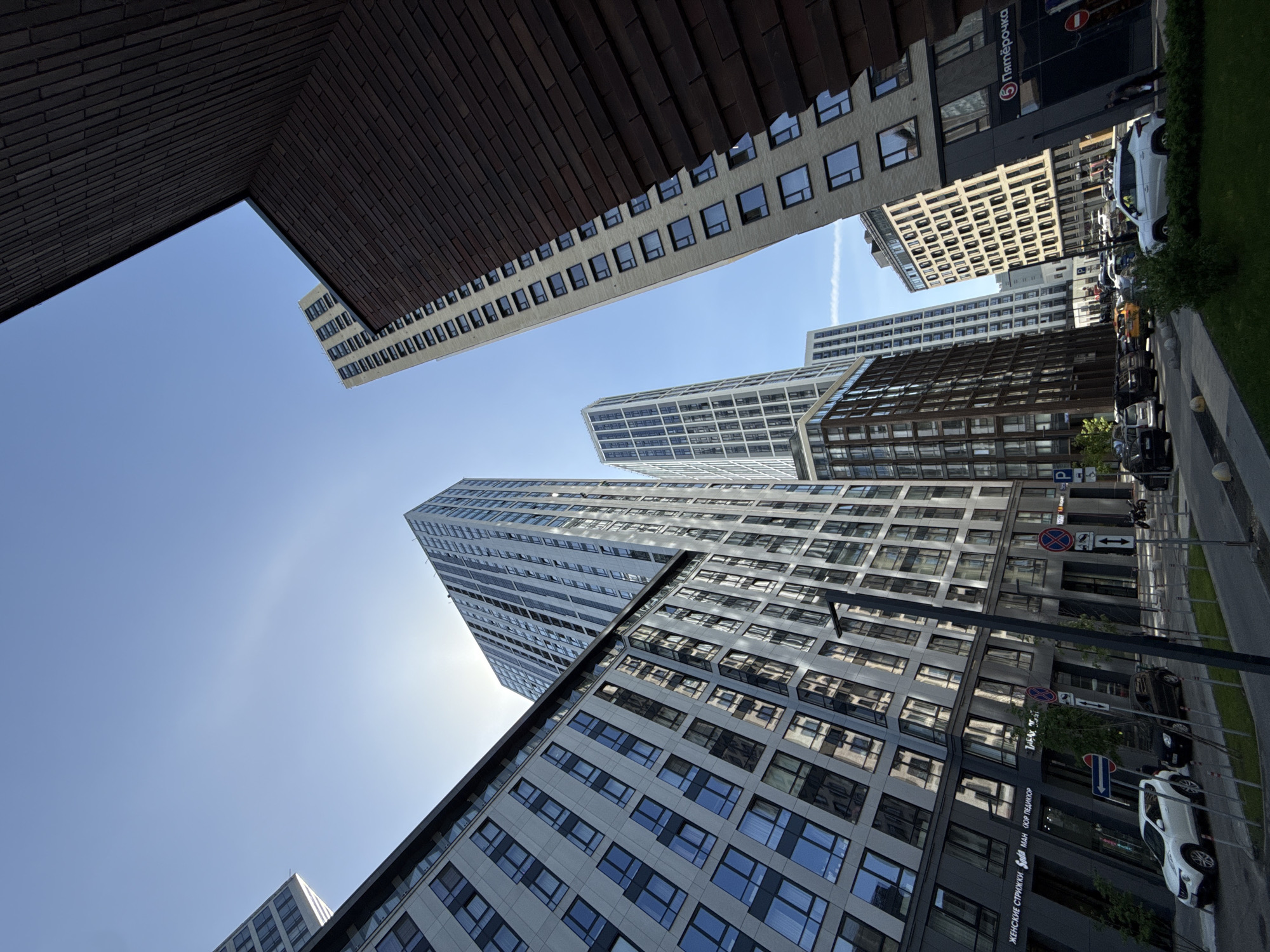 The image captures a low-angle view of a cluster of modern high-rise buildings in Moscow, Russia, under a clear blue sky. The architecture is characterized by repetitive window patterns and diverse facade materials, including brick and glass. The buildings appear to be located along a street with visible pedestrian walkways, parked cars, and street signs. The presence of a retail establishment like "Пятерочка" (Pyaterochka) further suggests the location is in an urban commercial area.