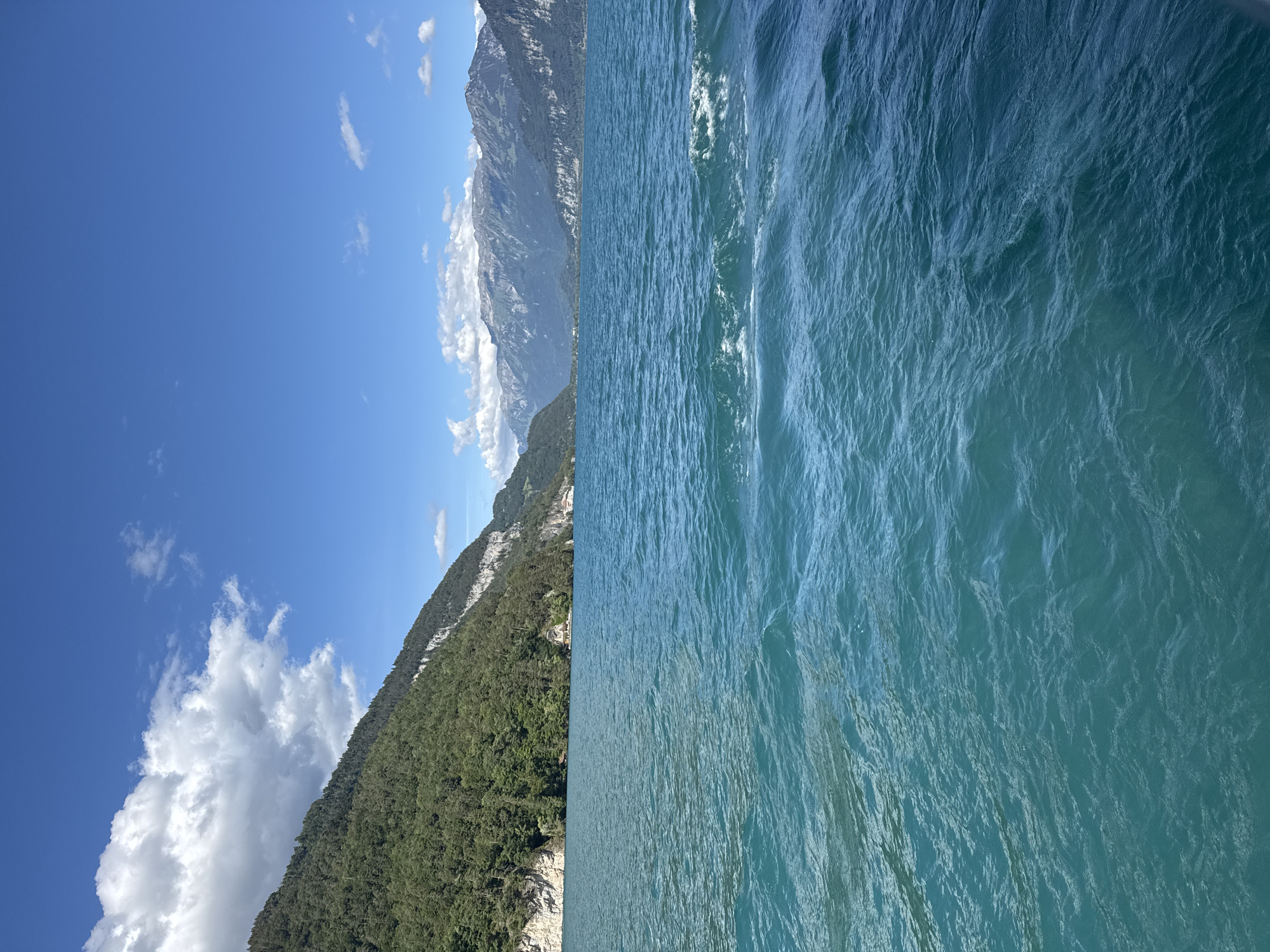 The image captures a serene view of Lake Thun from a boat, looking towards the mountains beyond Sigriswil, Switzerland. The foreground is dominated by the vibrant turquoise water of the lake, with gentle ripples and small waves suggesting movement. In the midground, a densely forested green mountain slopes steeply down to the water's edge, with a small village nestled along the shoreline, identifiable by scattered buildings and structures. The background features a majestic range of rugged, grey mountains under a clear blue sky dotted with fluffy white clouds. The overall impression is one of natural beauty and tranquility.