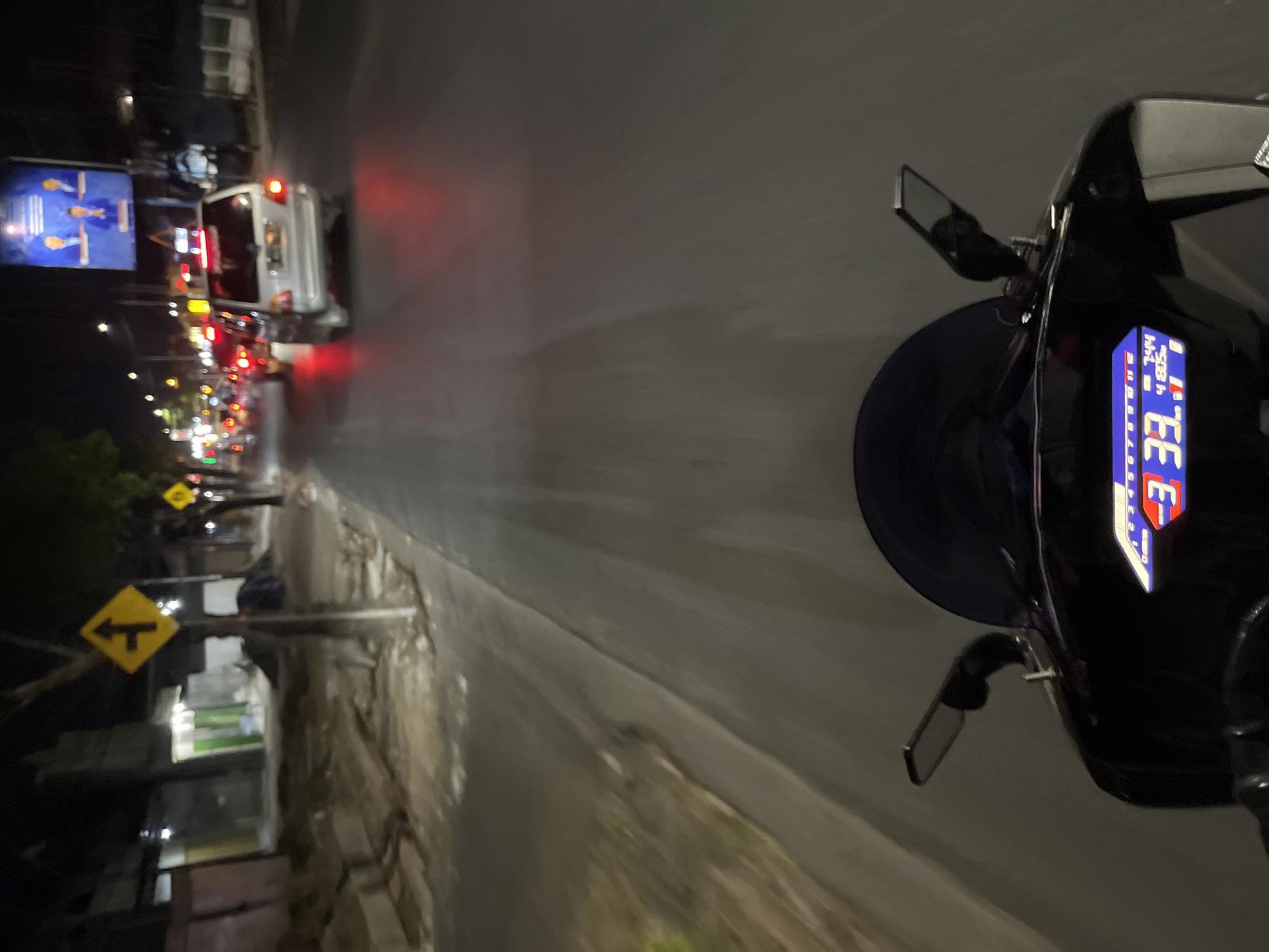 The image depicts a first-person perspective of riding a motorcycle at night on a paved road in Pecangaan, Indonesia. The rider's dashboard is visible in the foreground, showing a speed of 33 km/h and a fuel gauge indicating approximately half-full. A dark windshield and a motorcycle mirror are also prominent. The road is illuminated by streetlights and the headlights and taillights of other vehicles. In the background, a white SUV is visible in traffic, as are the red taillights of other cars. A billboard featuring three men in suits is mounted on a pole. To the left of the road, a sidewalk is under construction, with piles of dirt and construction materials. A yellow diamond-shaped road sign with a black arrow indicating a turn to the right and straight ahead is mounted on a pole. The overall environment suggests an urban or semi-urban setting at night.