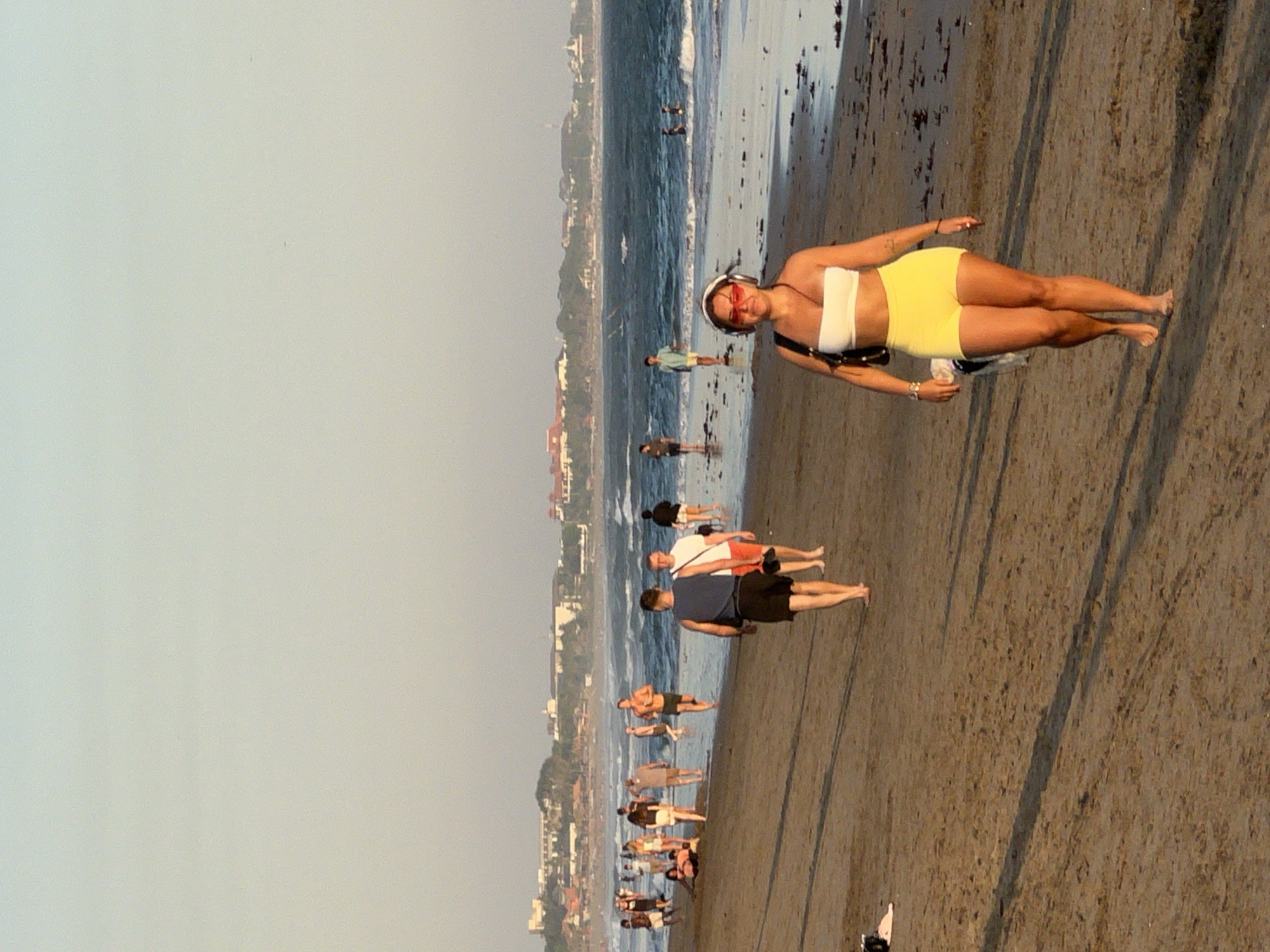 This is a full-body, eye-level shot of a woman walking on a sandy beach in North Kuta, Indonesia. The woman is in the foreground, facing the camera, wearing a white bandeau top and yellow shorts. She has red-rimmed sunglasses on and headphones over her ears. A black purse is slung over her right shoulder. She is barefoot and walking on the wet sand near the water's edge. Behind her, other beachgoers are visible walking along the shore and in the shallow water. In the background, the ocean is calm with gentle waves, and a distant coastline with buildings and palm trees is visible under a hazy sky. The overall lighting suggests it is late afternoon or early evening.
