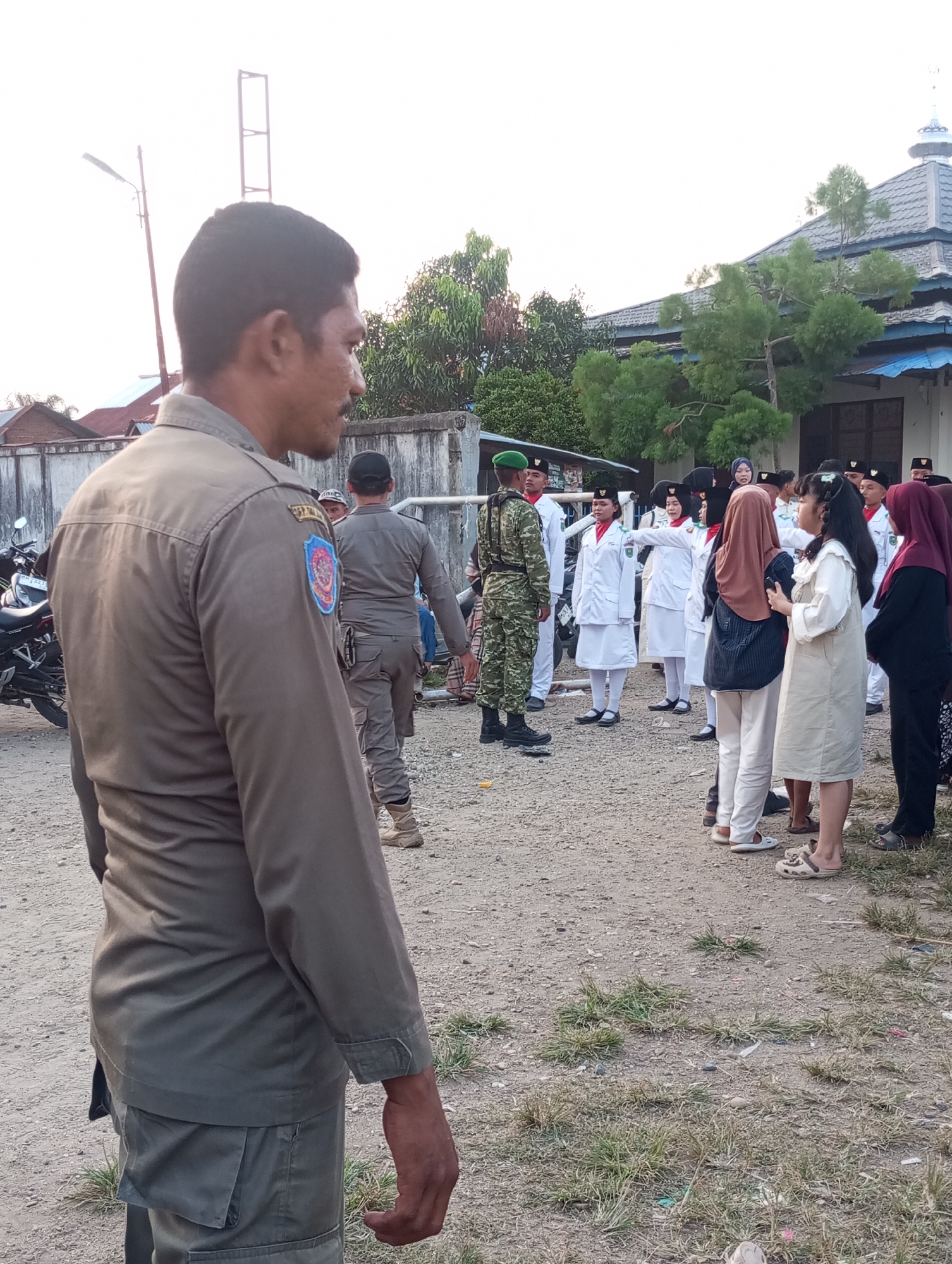 The image depicts an outdoor gathering in Candirejo, Indonesia. In the foreground, a man wearing a khaki uniform with a patch on his left shoulder stands facing away from the camera, his right arm slightly bent. Behind him, a group of individuals in white uniforms, some with red sashes, are arranged in formation. One individual in a camouflage uniform stands facing this group. Several people in casual clothing, including women wearing headscarves, are also present. A motorcycle is visible to the left of the frame, and a building with a tiled roof and a dome-like structure stands in the background. The ground appears to be dirt with patches of dry grass. The sky is bright, suggesting it is daytime.