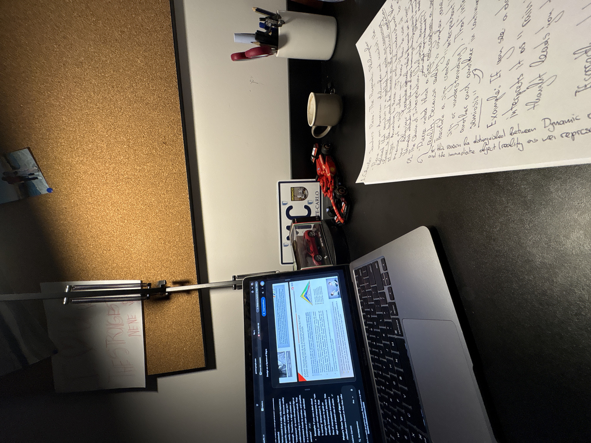 The image shows a desk with various items on it, oriented at an angle. On the left, there is a corkboard with a black frame, on which a note with the words "THE STRIGE NENE" is clipped. Next to the corkboard is a white wall, adjacent to a black desk surface. On the desk, there is a pen holder with pens and scissors, a small ceramic cup, a handwritten page with notes on Charles Sanders Pierce and semiotics, and a red Ferrari model car next to a "MONACO, ATE-CARLO" plate. A laptop is positioned on the right side of the desk, displaying a document about translation and semiotics. The laptop keyboard is visible. The background is a dark wall and what appears to be a window with translucent blinds. The scene does not contain any visual information to indicate the location is Novate Milanese, Italy.