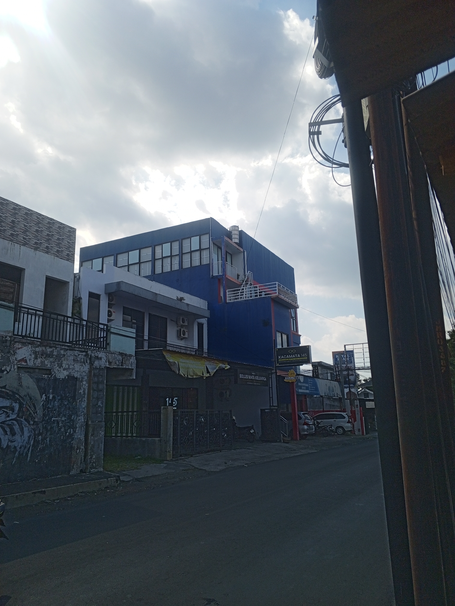 This media file captures a street scene in Jember, Indonesia, likely in the late afternoon or early evening, given the soft, diffused sunlight filtering through scattered clouds. The sky is a mix of bright white and grey clouds, suggesting a partly cloudy day.
The primary subjects are the multi-story buildings lining the street. The most prominent is a blue building with numerous rectangular windows on its upper floors, suggesting it might be an office or residential complex. To its left, a lighter-colored building with a grey brick facade and balconies is visible. Several storefronts are located at the ground level of these buildings.
Objects in the scene include parked cars and motorcycles, a few outdoor air conditioning units mounted on the buildings, and what appear to be street lamps or utility poles with coiled wires. Fencing and railings are also visible along the front of the buildings.
One of the notable details is the weathered appearance of the leftmost building's lower section, which has peeling paint and graffiti-like markings. This juxtaposition of modern and aging architecture creates a sense of history and lived experience in the location.
Visible text includes signs on the buildings. One sign reads "KACAMATA 145," indicating an optician or eyewear store. Another sign, partially obscured, seems to say "SOLUSI MATA KELUARGA." A street number "145" is clearly visible on a pillar in front of a gate. Other signs are too small or blurry to decipher. The overall mood of the scene is quiet and ordinary, depicting a typical urban street in Indonesia.