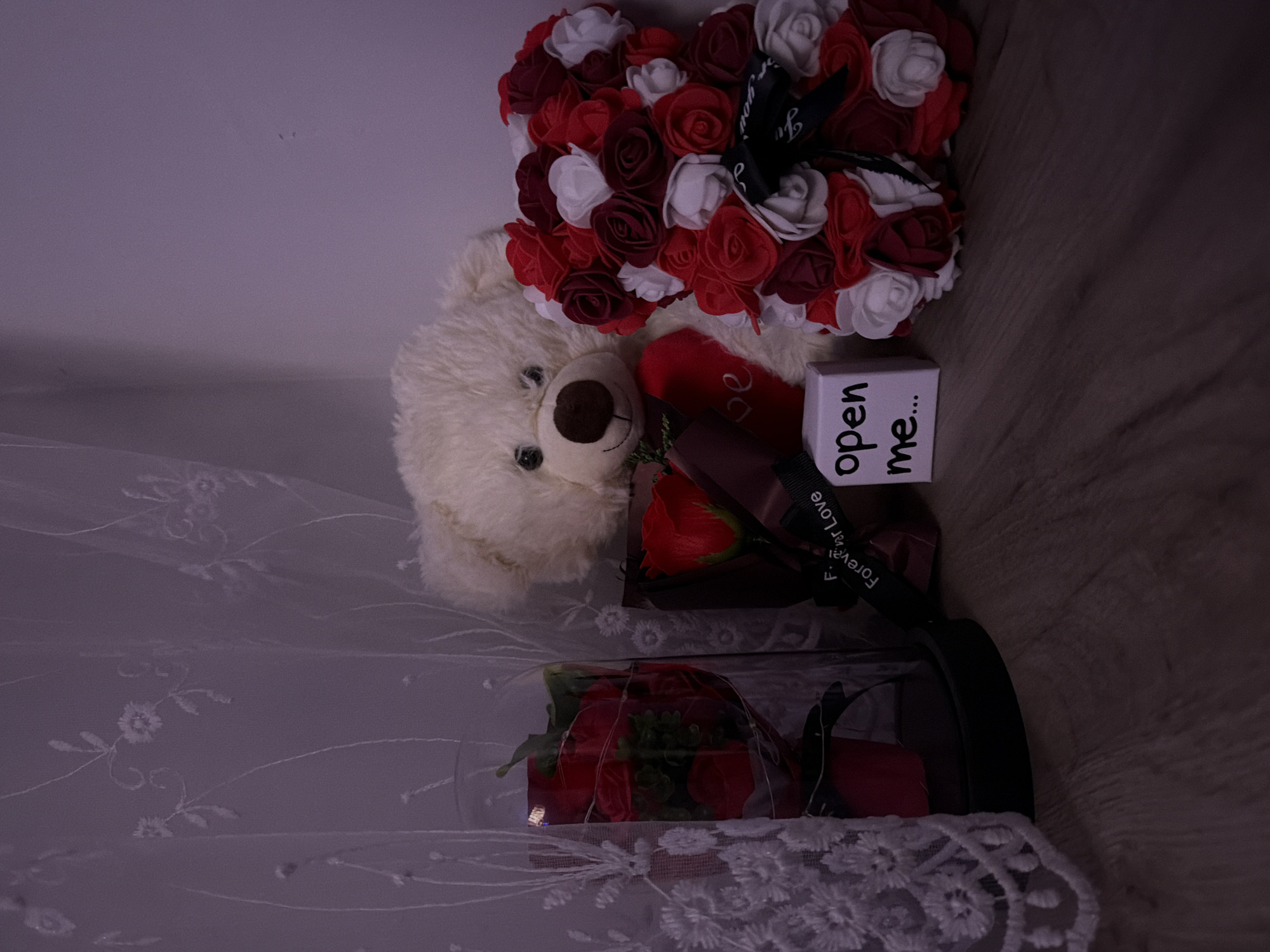 The scene, located in Varna, Bulgaria, depicts an indoor arrangement of decorative items on a light wood-patterned floor against a light-colored wall and a sheer, embroidered white curtain. On the far left, a glass cloche with a black base encloses multiple red artificial roses intertwined with string lights. Adjacent to it, a single red artificial rose is presented in a brown wrapper with a black ribbon partially reading "Forever Love." Centrally positioned is a large, white fluffy teddy bear with a brown nose and black eyes, adorned with a red heart-shaped item bearing the partial text "Love." To its right, a small white rectangular box is visible, labeled with the black text "open me..." Further right, a larger bear-shaped arrangement is constructed from numerous artificial red (dark and bright) and white roses, featuring a black ribbon around its neck with text partially visible as "...For You." The overall lighting is dim. No individuals are present.