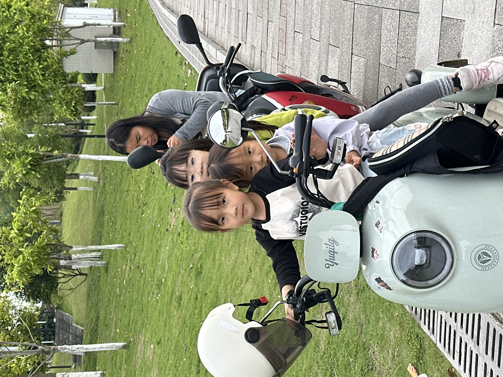 Five children are grouped together on and around two parked motor scooters on a paved surface bordering a grassy area with trees. The nearest scooter is light green with a white helmet hanging from the handlebar; the word "Yuqilg" is printed on the side. Behind it is a red and black scooter. A girl in a black and white shirt is in the foreground on the green scooter. Behind her, two younger girls and another older girl are on the bikes. A fifth person, in gray, is partially visible behind the second scooter. The location may be in the city of Jiangjin, China, as hinted in the prompt.
