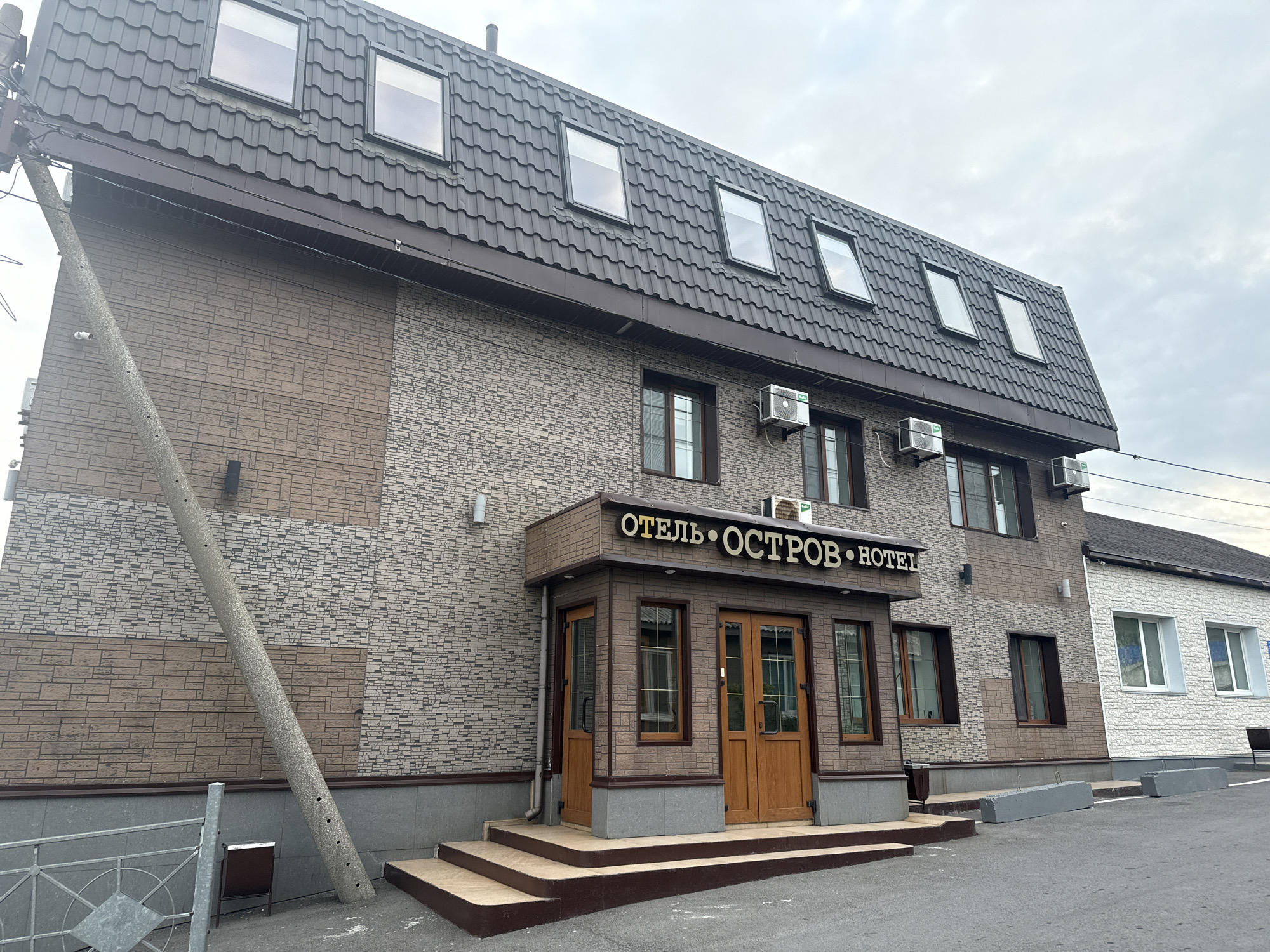 The image shows a two-story hotel building with a dark gray, textured roof. The facade of the building is clad in a mix of light gray and brown brick-like tiles. There are several dormer windows on the upper floor. The entrance to the hotel is marked by a brown awning with the text "ОТЕЛЬ • ОСТРОВ • HOTEL" in gold lettering. The entrance features two wooden doors and several windows. A set of concrete stairs leads up to the entrance. To the left of the entrance, a leaning concrete utility pole is visible, with a security camera mounted on it. In front of the building, there is an asphalt surface with some concrete barriers. To the right of the main building, a section of a white-clad structure with windows is visible. The sky is overcast. The scene is located in the city of Kuril'sk, Russia.