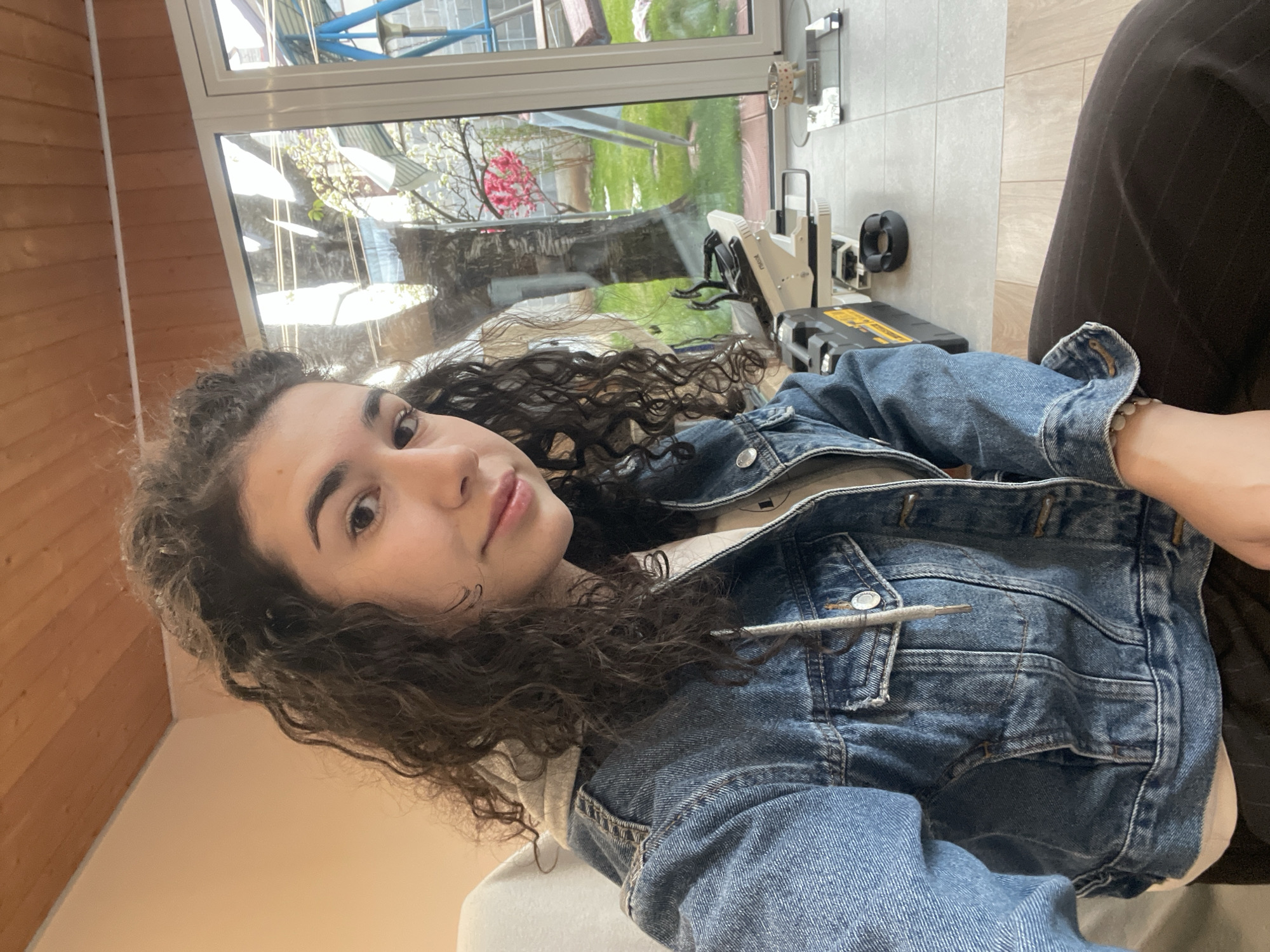 A young woman with curly dark hair is sitting indoors, wearing a denim jacket. A gray pencil rests in the jacket pocket. Outside the window, which is directly behind and above the woman, there's a yard with grass, trees, and a pink flowering bush. Reflections in the window show some sort of metal structure. Visible in the foreground, near the window, are tools and a toolbox on a tiled floor. The indoor wall is painted a light peach color. The woman is looking directly into the camera. It is not possible to ascertain the location in Sofia, Bulgaria, from this image.