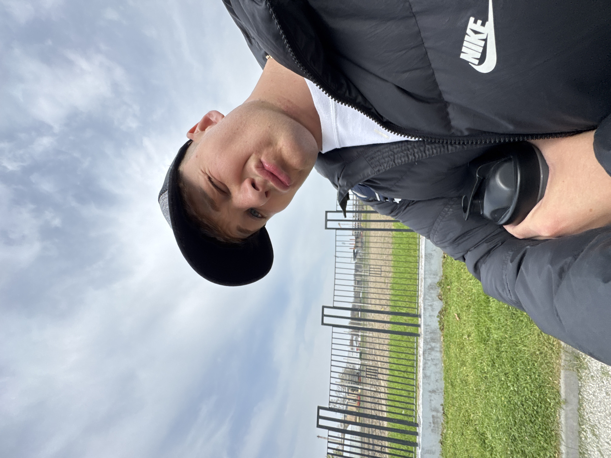 A man is photographed in an outdoor setting. He is wearing a black Nike jacket, a white t-shirt, and a black cap. He is holding a black shaker bottle in his right hand. The background includes a fence, grass, and a cloudy sky. Although the location is Plovdiv, Bulgaria, this cannot be confirmed from the image.