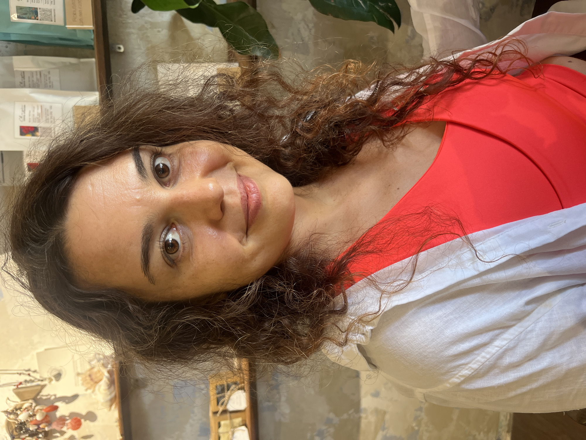 A woman is photographed from the chest up, looking directly at the camera with a slight smile. She has dark, curly hair that falls around her shoulders and a fair complexion with some freckles visible on her cheeks. She is wearing a bright coral-colored top under an unbuttoned, white linen shirt. The background is slightly blurred, showing shelves with various items, possibly in a shop. To the right, a large green leaf from a plant is visible. The lighting suggests an indoor setting, possibly in the city of Phu Thuy Ward, Vietnam, given the context.