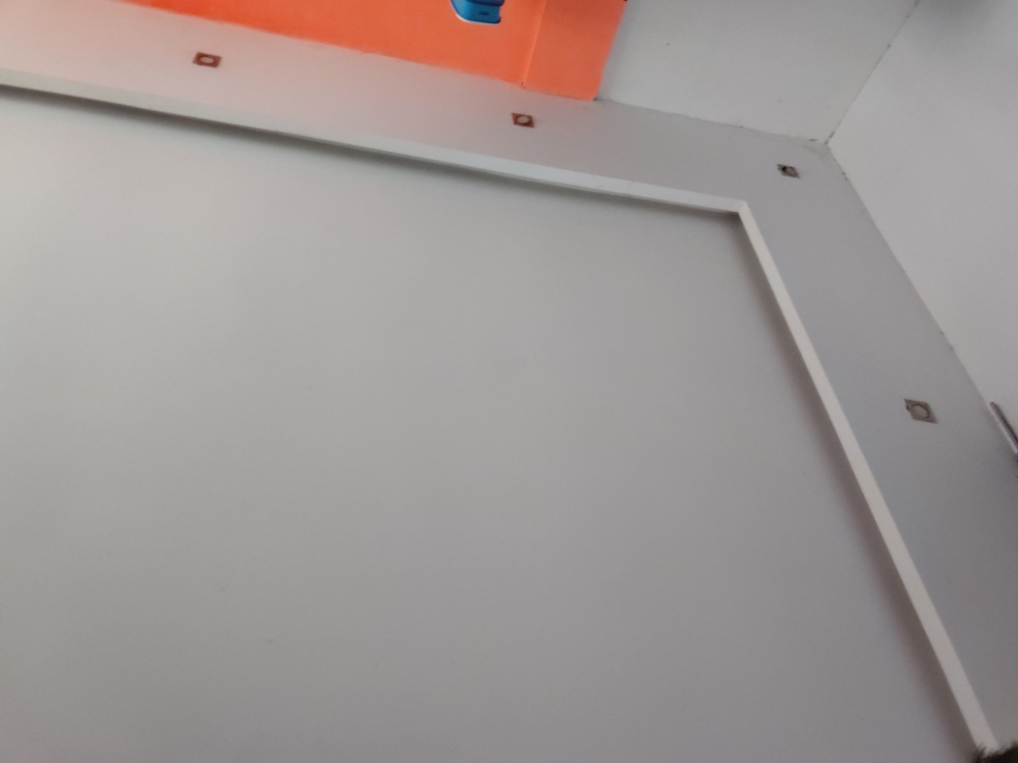 The image displays an indoor setting, focusing on a corner where light gray walls meet a white ceiling. A prominent feature is a large, light gray rectangular panel or framed section on the left wall, outlined by a clean white border. Above this panel, a distinct orange-colored surface or wall segment is visible, contrasting sharply with the lighter tones of the room.
Several small, dark, square-shaped openings or embedded inserts are visible on the white/light gray wall area, particularly along the top edge above the framed panel and in the upper right corner. These appear to be evenly spaced in some areas, suggesting they might be mounting points, outlets, or unfinished elements for fixtures. One of these openings reveals a dark, reddish-brown interior.
In a hint crop, a small blue object with a rounded top is partially seen on the orange surface, and a thin, metallic or silver object is visible against the right wall, near one of the square openings. At the very bottom right, some dark, possibly fibrous material is partially visible, which could be part of a cleaning tool or debris.
The scene is static, depicting a clean, possibly newly constructed or renovated space. There is no visible activity, people, or discernible text. The lighting suggests an indoor environment, but the time of day and weather are not indicated. The overall impression is one of modern or functional interior architecture.