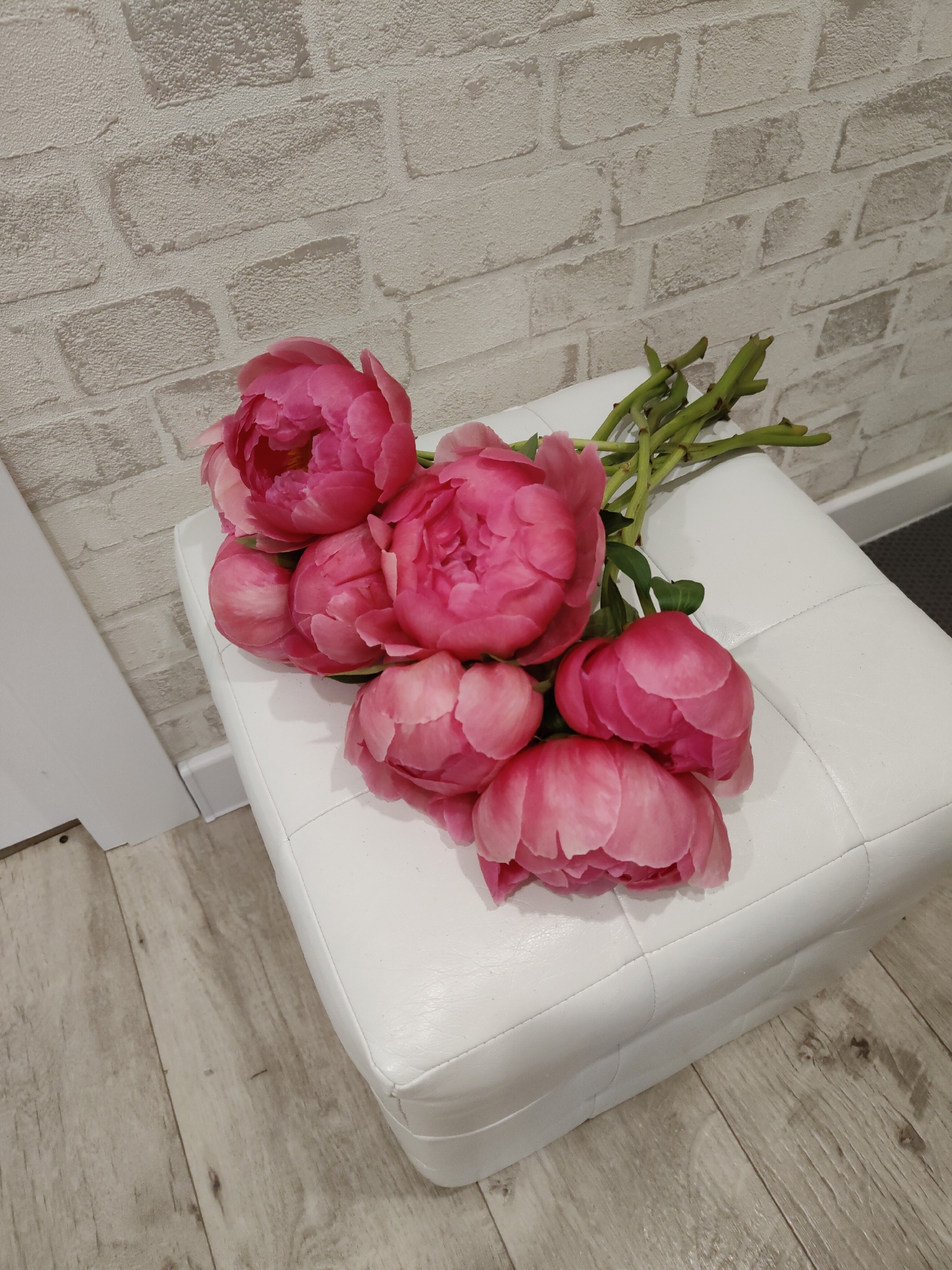 This image presents an indoor still life in the city of Unknown, Unknown. A cluster of seven pink peonies, displaying varying stages of bloom, is arranged horizontally on the white, square-tufted surface of an upholstered ottoman. The peony stems are visible and green. Behind the ottoman, a wall is covered with light-toned wallpaper designed to resemble irregular, textured brickwork. The floor beneath the ottoman consists of light-colored wooden planks exhibiting visible grain. A white baseboard runs along the bottom of the wall. To the left, a vertical white architectural element, likely a door frame, is partially visible. A dark, possibly grey or black, floor covering is partially visible on the far right, contrasting with the wooden floor. No individuals are present.