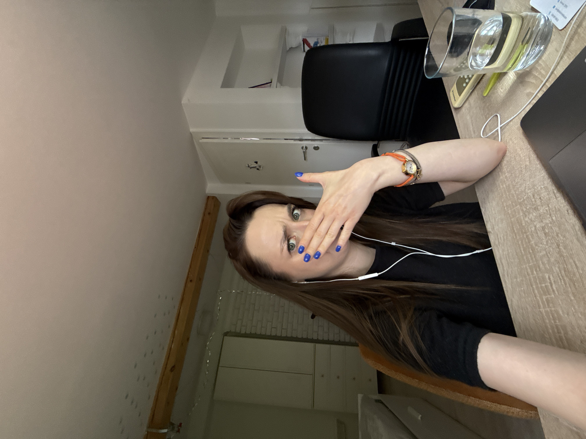 A young woman is captured in a selfie-style portrait, appearing rotated clockwise and looking directly at the camera with a wide-eyed expression of surprise or mild shock. She has long, dark brown hair and is wearing a black top. Her right hand, adorned with bright blue nail polish, is covering her mouth, with her index finger slightly extended. White wired earphones are visible around her neck and in her ears. On her left wrist, she wears a watch with an orange strap and a small, circular gold face.
She is seated at a light-colored wooden desk, where a clear glass filled with water, part of a dark laptop, and a beige electronic device are visible. A black office chair is behind her. The room features light-colored walls and a sloped ceiling, suggested by a wooden beam running along the upper left, hinting at an attic or converted space. In the background, white cabinets or shelves are present, and a section of the wall displays a subtle, light-colored brick pattern. The lighting indicates it is daytime indoors. The setting appears to be a home office or personal workspace within Palilula Urban Municipality, Serbia. No discernible text is clearly visible in the image.