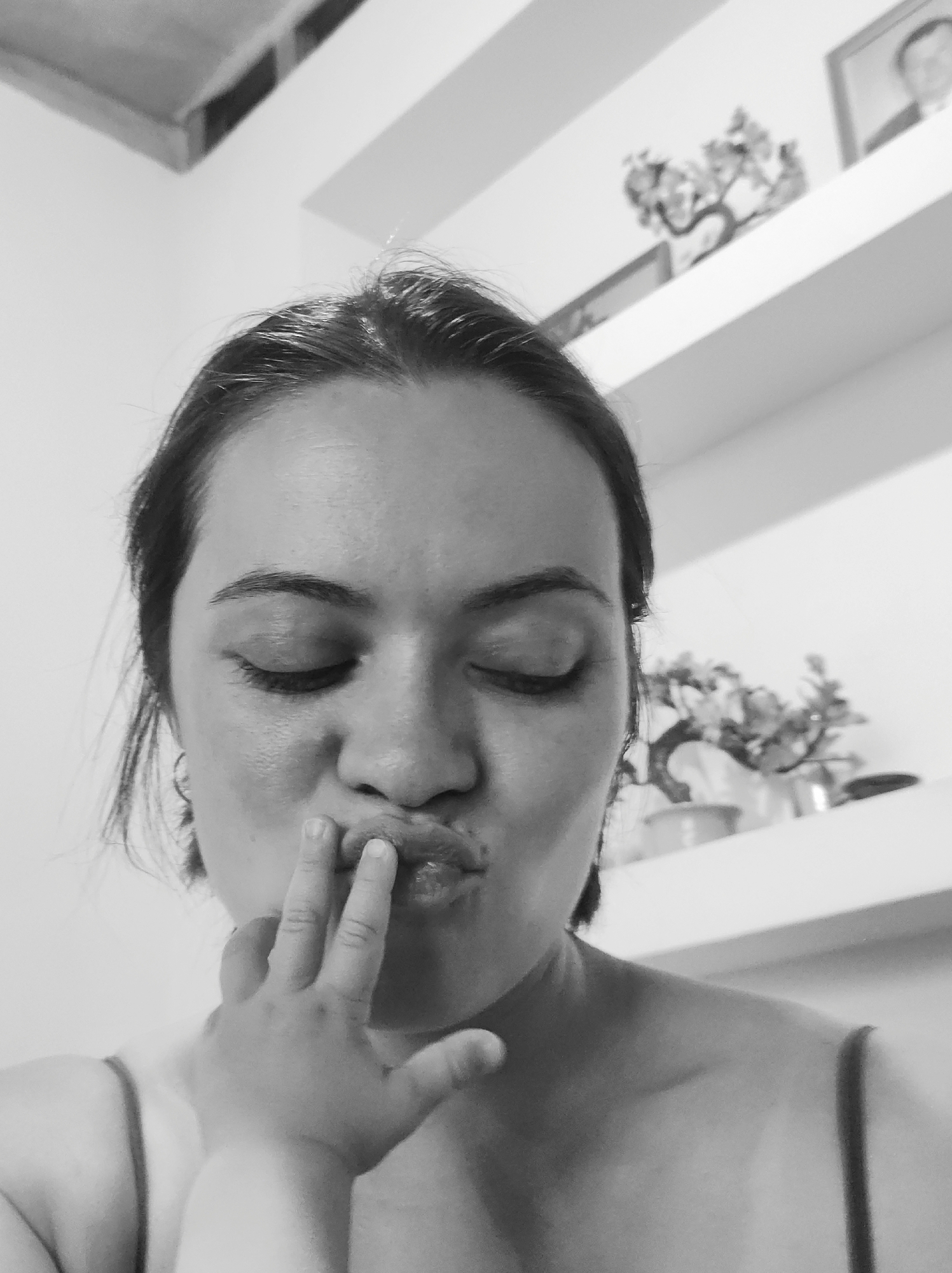 A black and white, close-up photograph captures a woman from the chest up, her eyes closed, with her lips pursed into an O-shape. A small, infant-sized hand with short fingernails has its index, middle, and ring fingers pressed against the woman's lips. The woman has dark hair pulled back from her face and is wearing a thin-strapped top. The background is a stark white wall featuring multiple horizontal shelves. On these shelves, a small, dark, artificial tree-like decoration and a partially visible framed picture containing a blurry image of a person are discernible. A dark rectangular object, possibly an air vent, is visible in the upper left quadrant of the image. The scene is illuminated by ambient light within an interior space in Unknown, Unknown.