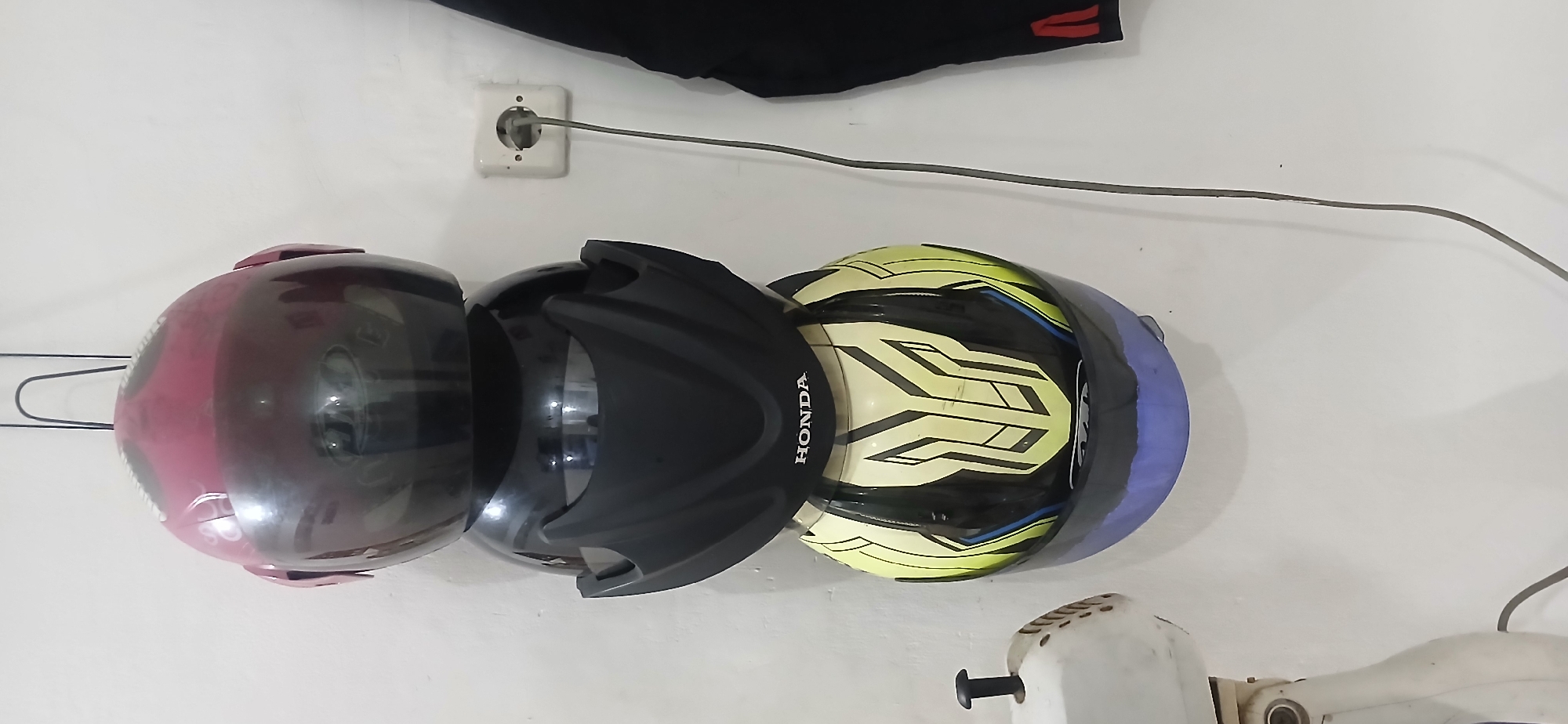 Four motorcycle helmets are stacked vertically on a white wall in a residential setting. The top helmet is pink with a dark visor. Below it is a black helmet. The third helmet from the top is black with a prominent "HONDA" logo on the front. The bottom helmet is yellow and black with a graphic design and a blue visor. To the right of the helmets, a white electrical outlet is visible with a gray cord plugged in. To the left of the helmets, a portion of a white object, possibly a fan, is visible. A black garment hangs on the far right side of the image. The scene is consistent with a home environment in West Jakarta, Indonesia, where motorcycle helmets are commonly stored.