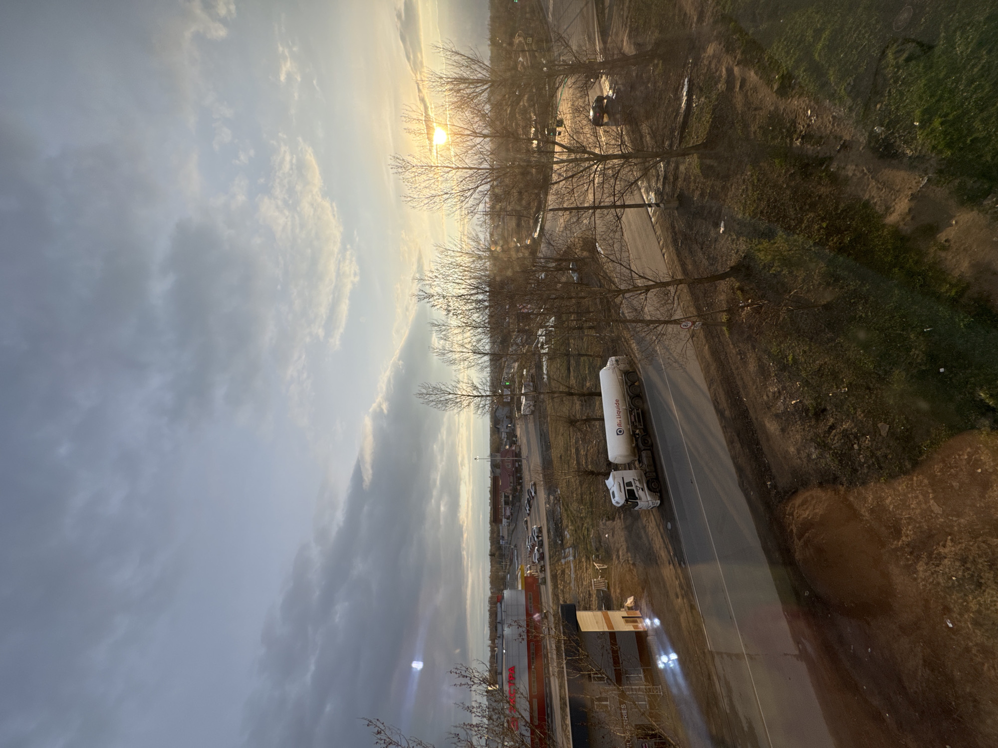 The image shows a view over Kladovitsy, Russia, featuring a cloudy sky with a sunburst effect peeking through bare trees. A road runs through the middle ground, where a white truck with a cylindrical tank trailer is parked. Further along the road, there are parked cars and what seems to be commercial buildings, possibly a store, with a partially constructed structure at the lower edge. The immediate foreground is blurry, perhaps the inside of a window. The right side of the image shows a patch of grassy or barren land.