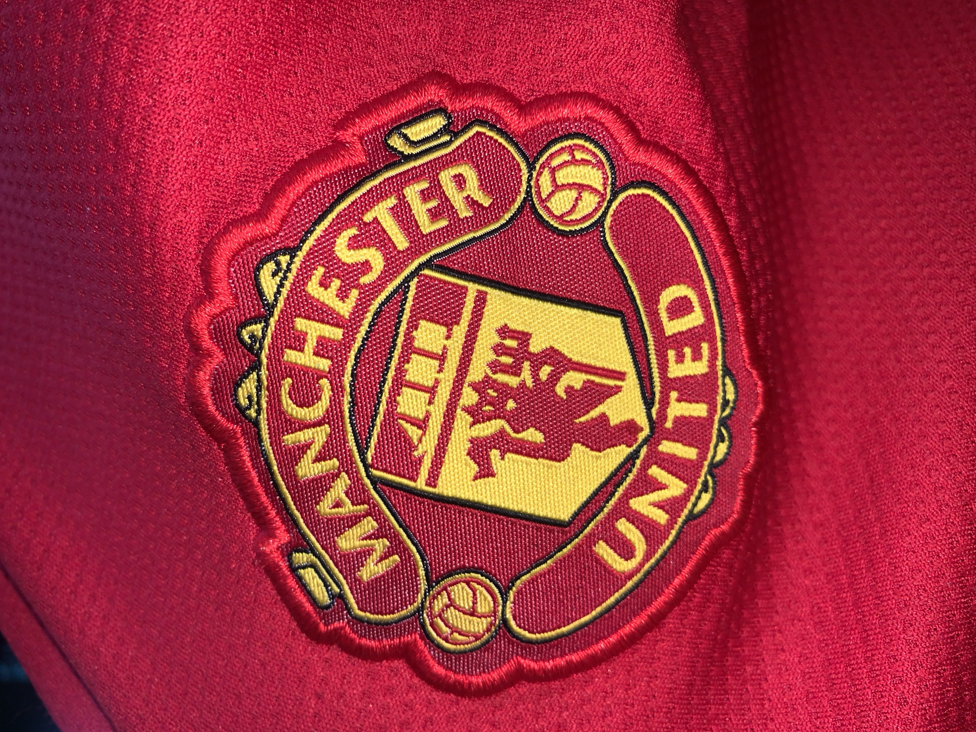 The image shows a close-up of the Manchester United football club emblem embroidered on a red fabric, possibly a jersey. The emblem features the words "Manchester United" arched above and below a shield with a devil on it. Two footballs are situated in the emblem. No location is visible. The image is cropped in a manner that does not show any surroundings or individuals, nor anything related to the stadium in Nigeria.