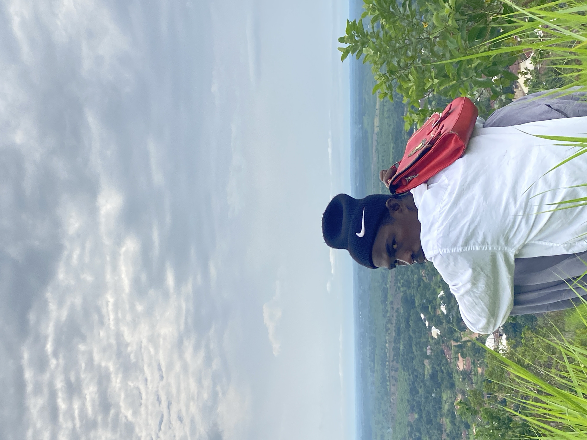 A young man wearing a black beanie with a white Nike logo and a white long-sleeved shirt is sitting on a grassy slope overlooking a landscape. He is holding a red handbag over his shoulder. The background shows a hazy view of a town with scattered buildings and lush green vegetation, characteristic of the area around Ufu Esioni, Nigeria. The sky is overcast with layered clouds. Tall, green grass is in the foreground, partially obscuring the lower part of the man's body.