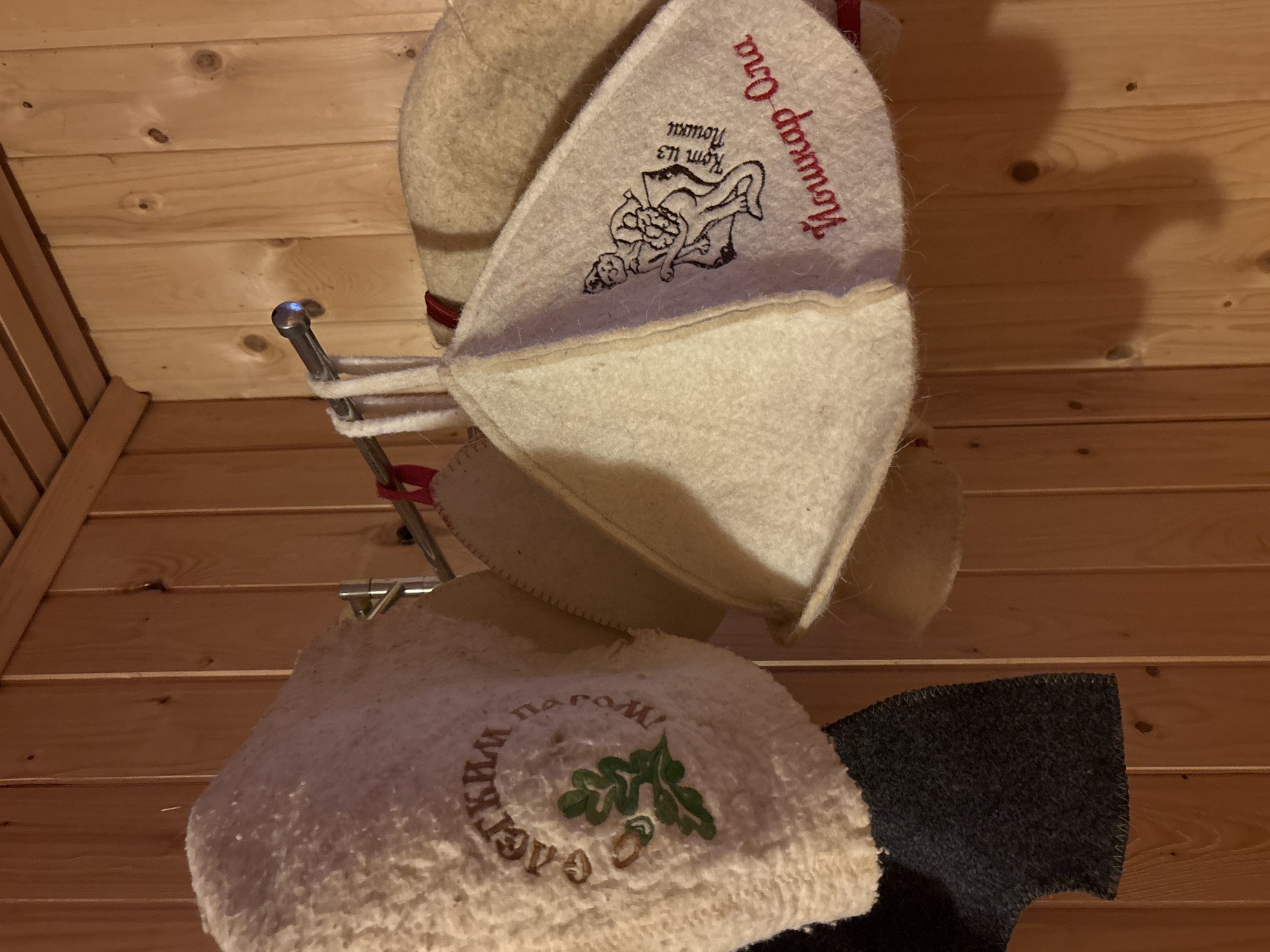 The image shows a collection of bath hats hanging on a rack. The hats are made of felt and embroidered with designs. One hat has an image of a reclining figure and the words "Nuscap Ora" and "Kom uz nouxm" embroidered on it. Another hat has an embroidered design of oak leaves and acorns, surrounded by Cyrillic text. A dark gray rectangular object is next to the second hat. The background consists of wooden planks, likely a wall or ceiling. The location is Prikamskii, Russia.