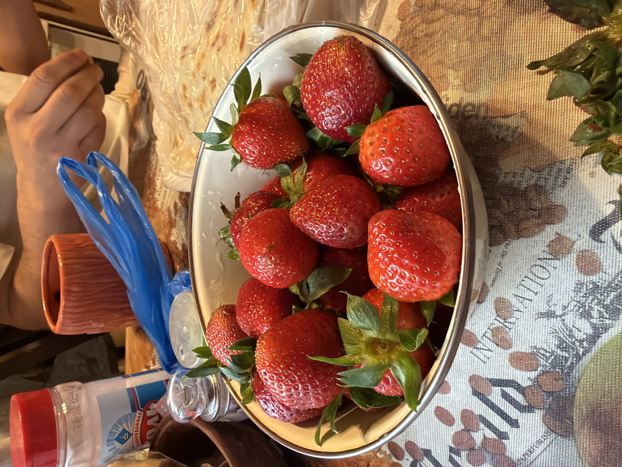 The image shows a bowl filled with strawberries. The bowl is oval-shaped, with a white interior and a metallic rim. The strawberries are red, ripe, and have green stems and leaves. The bowl sits on a table with a patterned tablecloth. Also present are what appears to be food in a clear plastic wrapping, a blue plastic bag, and an open glass bottle with a red-capped container next to it. In the background, there is a hand and part of an arm. Based on the container labeling "Моссоюзплодовощ" (Mossoyuzplodovosh), which translates to "Moscow Union of Fruit and Vegetables", the objects may be located in Moscow, Russia.