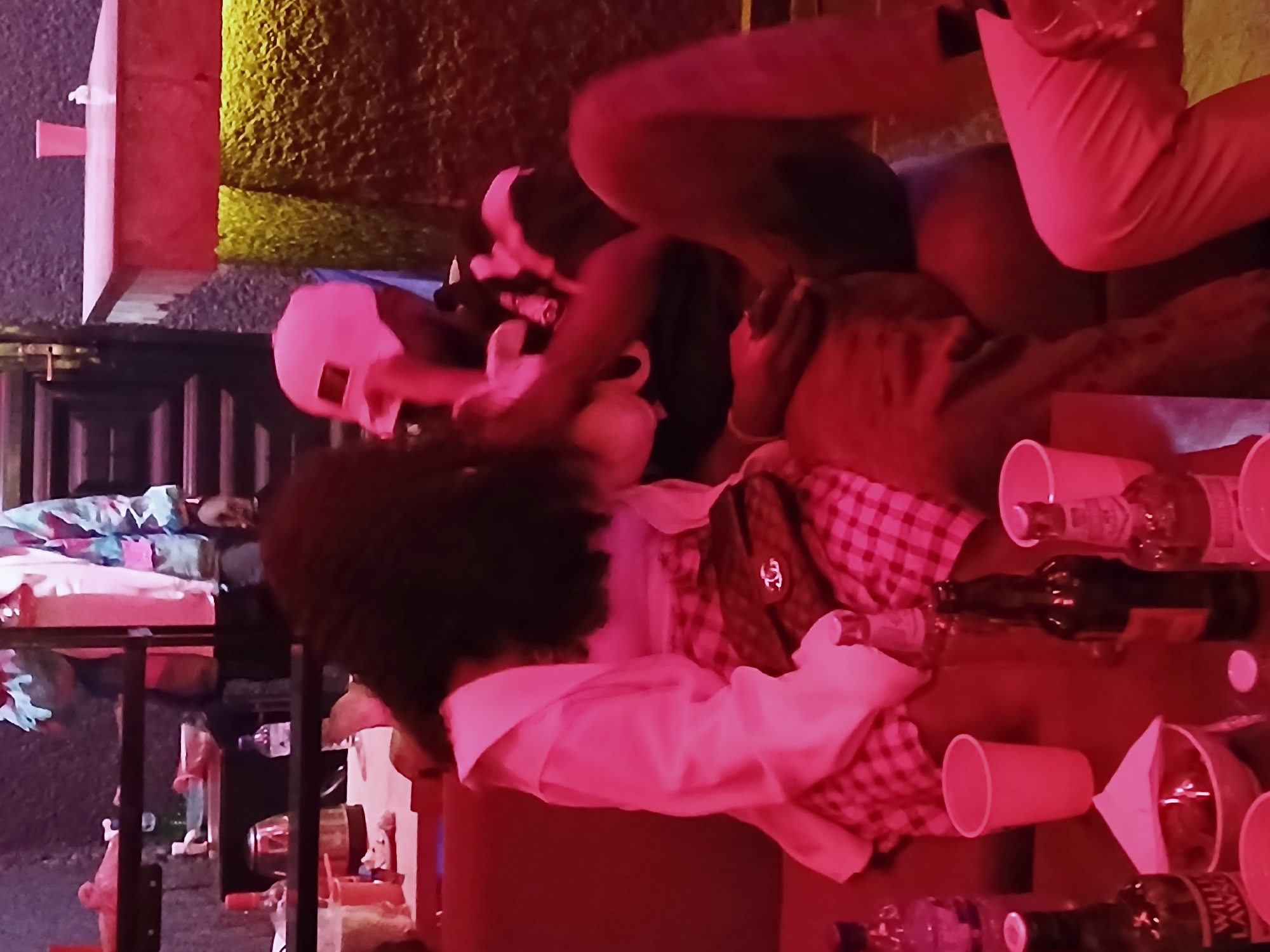 The image depicts a scene on Rukuba Road, Nigeria, showing several individuals engaged in social activity. There are bottles and cups scattered around, suggesting a gathering involving drinks. The overall lighting appears to be a reddish hue. One person has an afro and is wearing a checkered shirt and a white jacket. Other individuals can be seen, with one wearing a white hat and another pink pants. There are stairs and furniture visible in the background.