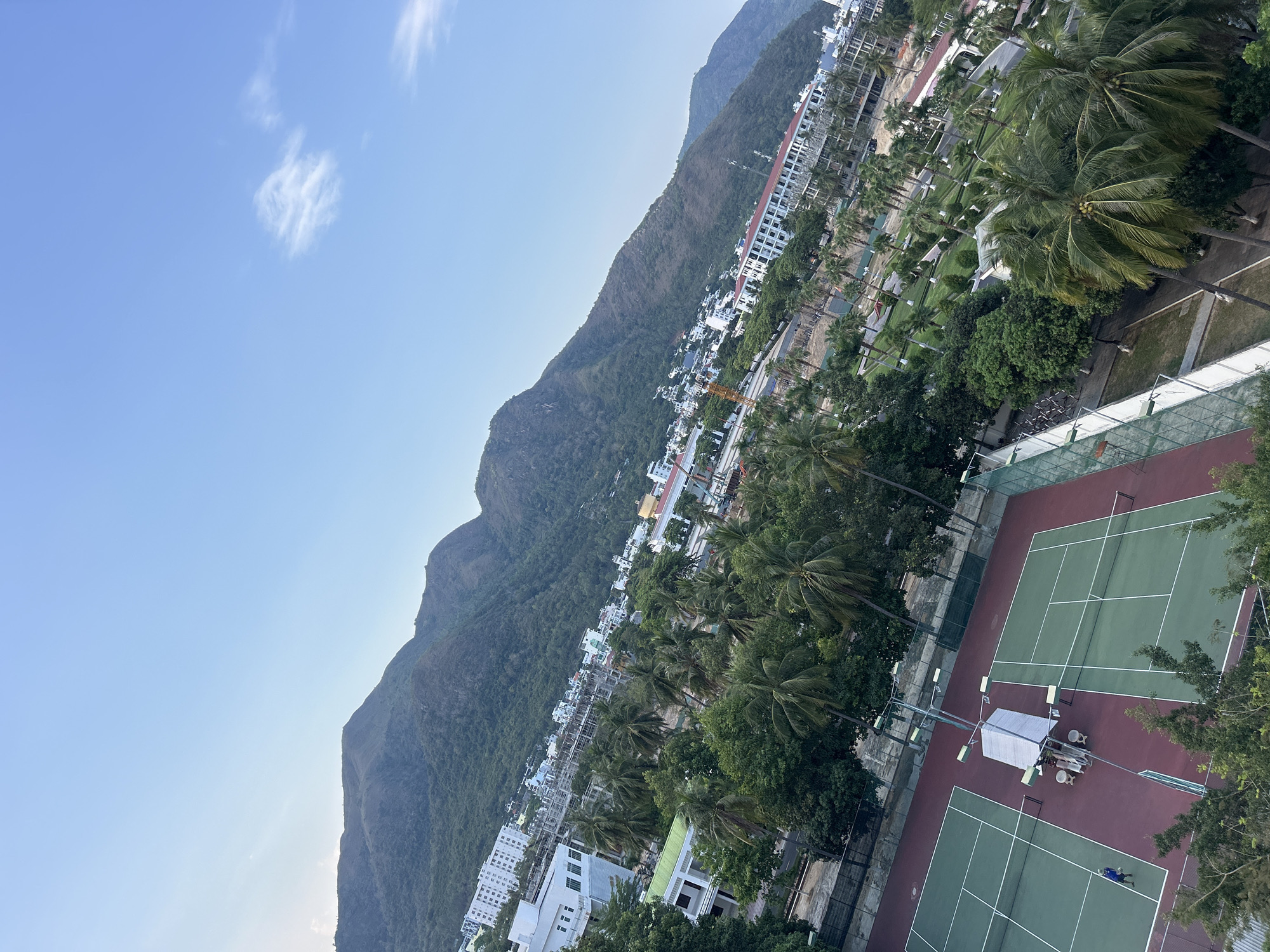This high-angle shot reveals the Bac Nha Trang Ward in Vietnam, showcasing the juxtaposition of urban development and natural landscapes. Mountains loom in the background, their green slopes providing a stark contrast to the city's architecture. Buildings, both complete and under construction, dot the landscape amidst lush trees and palm trees. In the foreground, tennis courts are visible. A person is visible on one of the courts.
