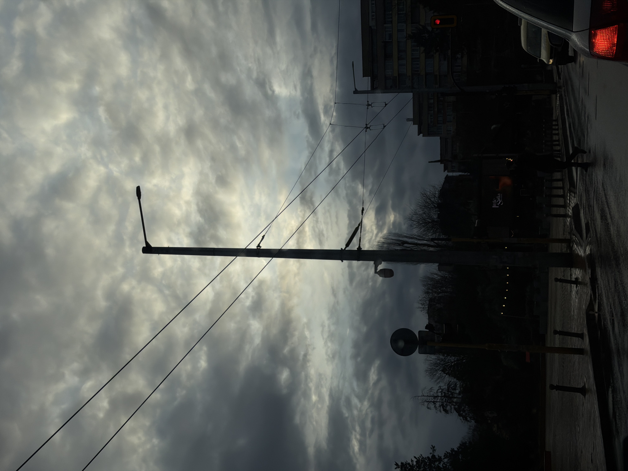 Captured from within a vehicle, the scene overlooks a street in Sofia, Bulgaria, marked by overcast skies. Street lamps stand in the foreground, connected by overhead wires. Traffic lights are visible, as well as buildings in the background. A pedestrian is walking on the street. The weather appears dark and cloudy.