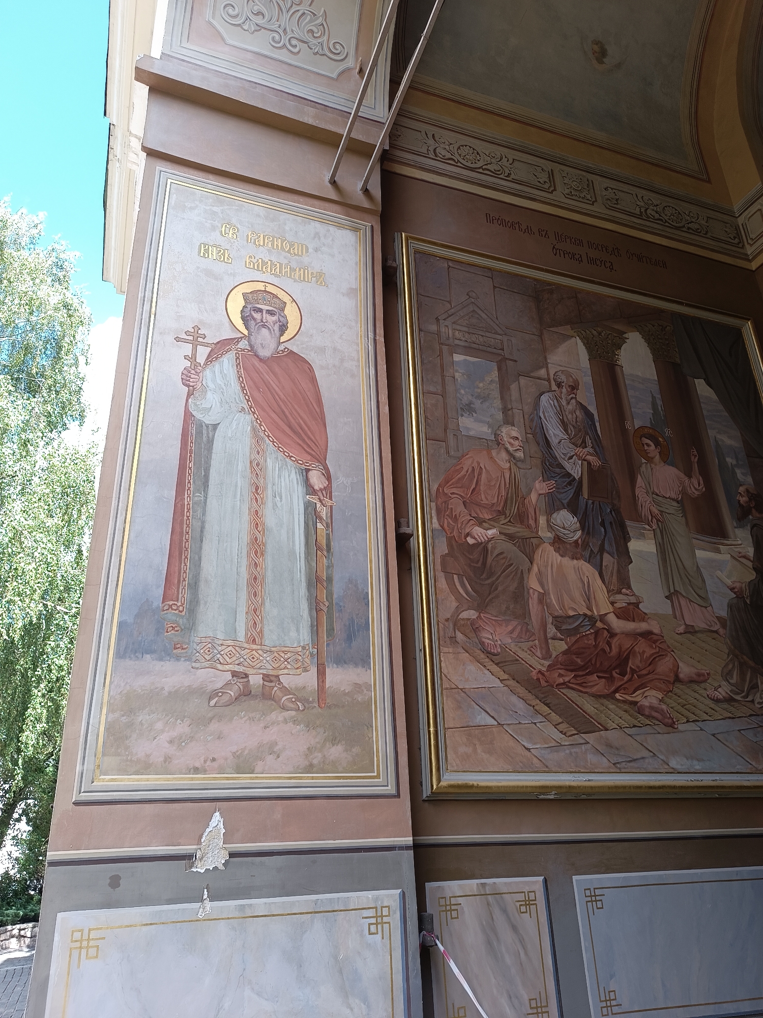 A fresco depicting religious figures adorns an exterior wall. On the left, a stylized image shows a bearded man with a halo and crown, holding a cross in his left hand and a sword in his right. He wears a red cloak over a light blue robe with ornate trim. Above him, text is inscribed in gold Cyrillic script. To the right, a larger fresco depicts a scene with several men, likely apostles, in flowing robes. A central figure, possibly Jesus, with a halo, stands gesturing with his right hand. The setting appears to be an architectural interior with columns. The overall fresco is framed by decorative borders. The wall surface below the frescoes shows signs of damage with peeling paint.