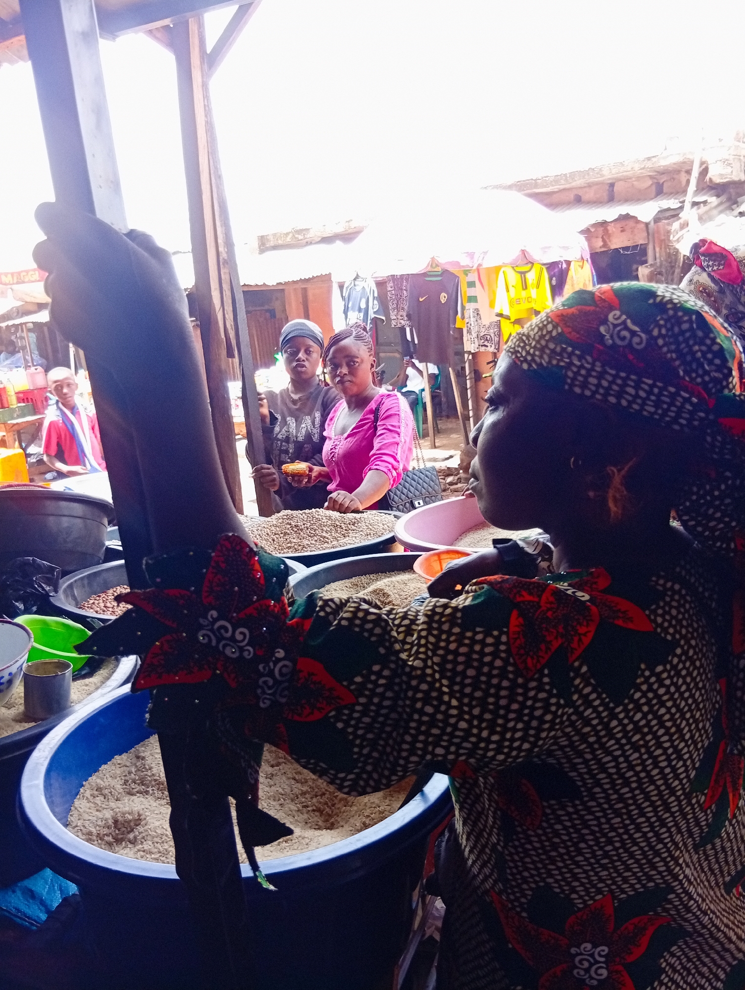 In Bukuru, Nigeria, a woman wearing a headscarf and a patterned dress stands behind a market stall filled with bags of grains. Her arms are extended, and she appears to be interacting with a customer who is holding a small amount of grain in their hand. Two other individuals are visible behind the woman, looking towards the camera. The market stall has overhead coverings, and various items, including clothing, are displayed in the background. The overall setting is an outdoor market with a mix of natural light and artificial lighting.