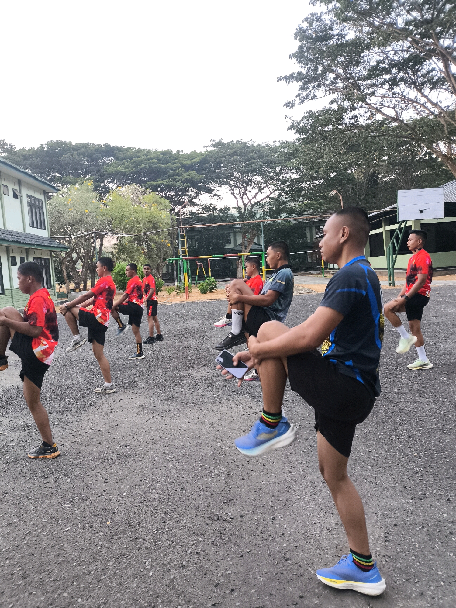 In a paved outdoor area with gravel, several young men in athletic wear are engaged in a group exercise routine. The individuals are wearing matching red t-shirts with a black and white pattern, or solid dark blue shirts, and black shorts. They are performing leg stretches, with some holding one knee towards their chest while standing on the other leg. In the background, there is a light green building with dark windows, trees with dense foliage, and a basketball hoop with a backboard mounted on a green metal frame. The ground is a mix of asphalt and gravel. The lighting suggests it is daytime, possibly early morning or late afternoon.