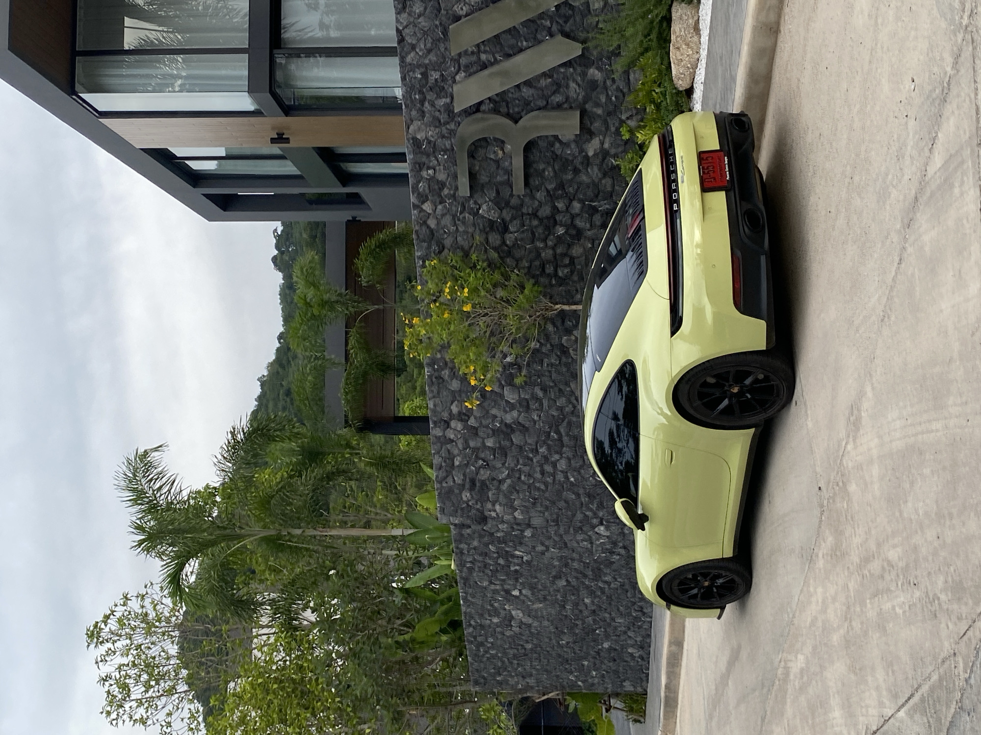 The image depicts a light-yellow Porsche parked next to a building with a stone facade. A sign is mounted on the facade. The building appears to be a modern architectural structure with multiple floors and large windows, partially visible. Lush greenery, including various trees and shrubs, surrounds the building and extends into the background. The image indicates the location in the city of Choeng Thale, Thailand, is suggested by the license plate of the Porsche which reads "ป-4515", a car plate identifier in Thailand. The scene appears to be taken during daylight under an overcast sky. The ground is paved with concrete.