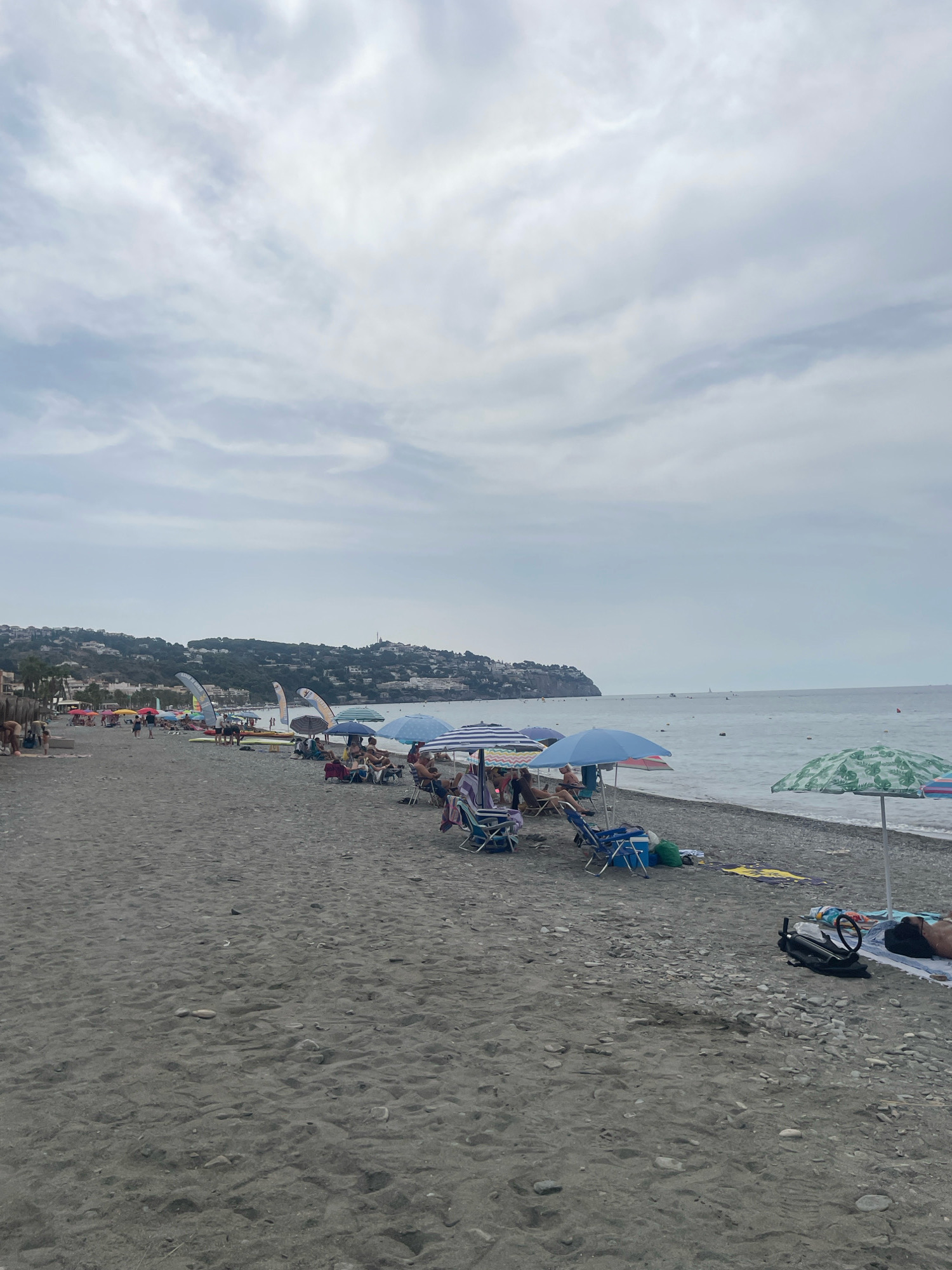 This is a medium shot of a crowded pebble beach in La Herradura, Spain, on an overcast day. The beach is lined with rows of colorful umbrellas and lounge chairs, with people sitting and relaxing. In the background, the Mediterranean Sea is visible, with a few boats in the distance. The coastline is lined with buildings and lush green hills. Several individuals are seen walking along the beach or sitting under the umbrellas. Some water sports equipment, including surfboards and windsurfing sails, are also visible.