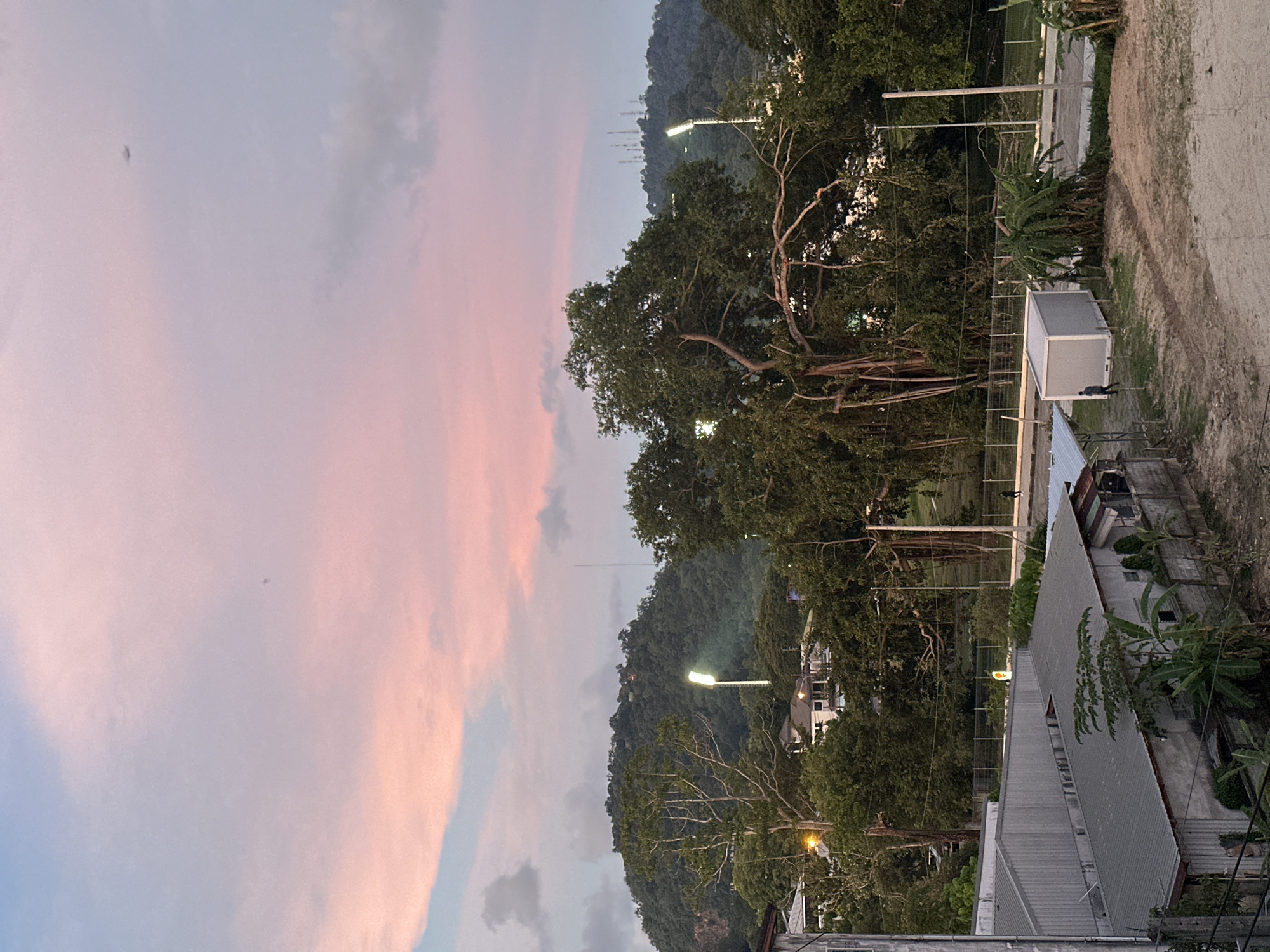 The image depicts a sunset scene in Wichit, Thailand. The sky is a gradient of pink and blue, with scattered clouds. In the foreground, there are several buildings with corrugated metal roofs, and some vegetation. In the middle ground, there are trees, a road, and streetlights. In the background, a lush green hill is visible, with more trees and some structures. An airplane is flying in the sky.