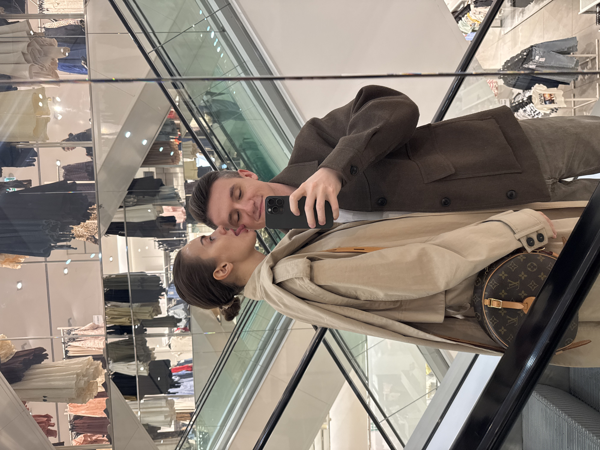 A couple is kissing in front of a mirror. The man is wearing a brown jacket and holding a phone to take a picture of them. The woman is wearing a tan trench coat and a Louis Vuitton bag. They appear to be in a store, as clothes racks are visible in the mirror. They are standing on an escalator. The location is Unknown, Unknown.