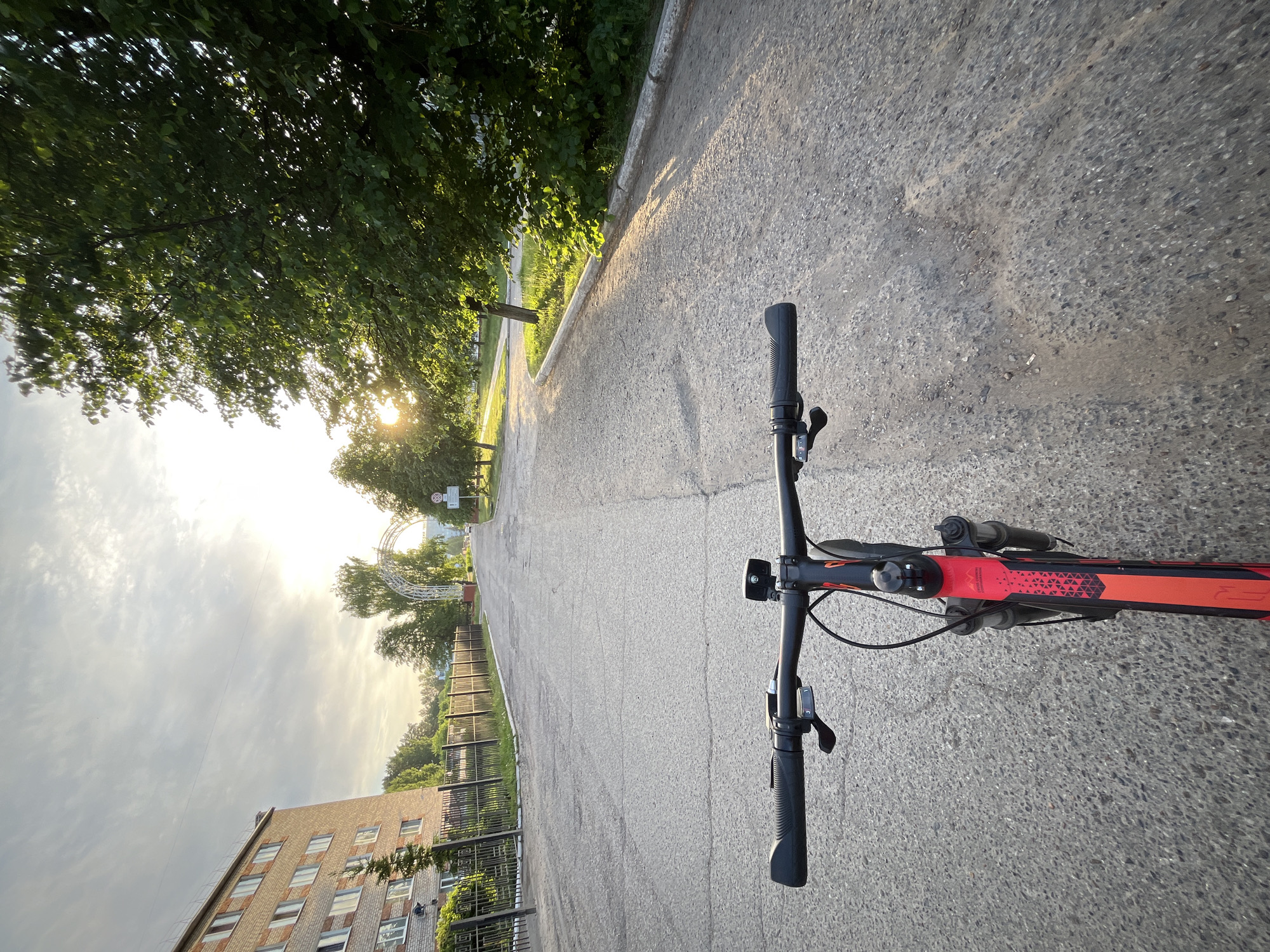 View from the perspective of someone riding a bicycle on a paved road. The bicycle handlebars and part of the frame, which is red and black, are visible. The road stretches straight ahead towards an arched gateway structure in the distance, partially obscured by trees. To the left, there is a building with multiple windows and a black metal fence. Trees line both sides of the road. The sun is low in the sky, creating bright glare. The scene appears to be in the city of Chaikovsky, Russia.