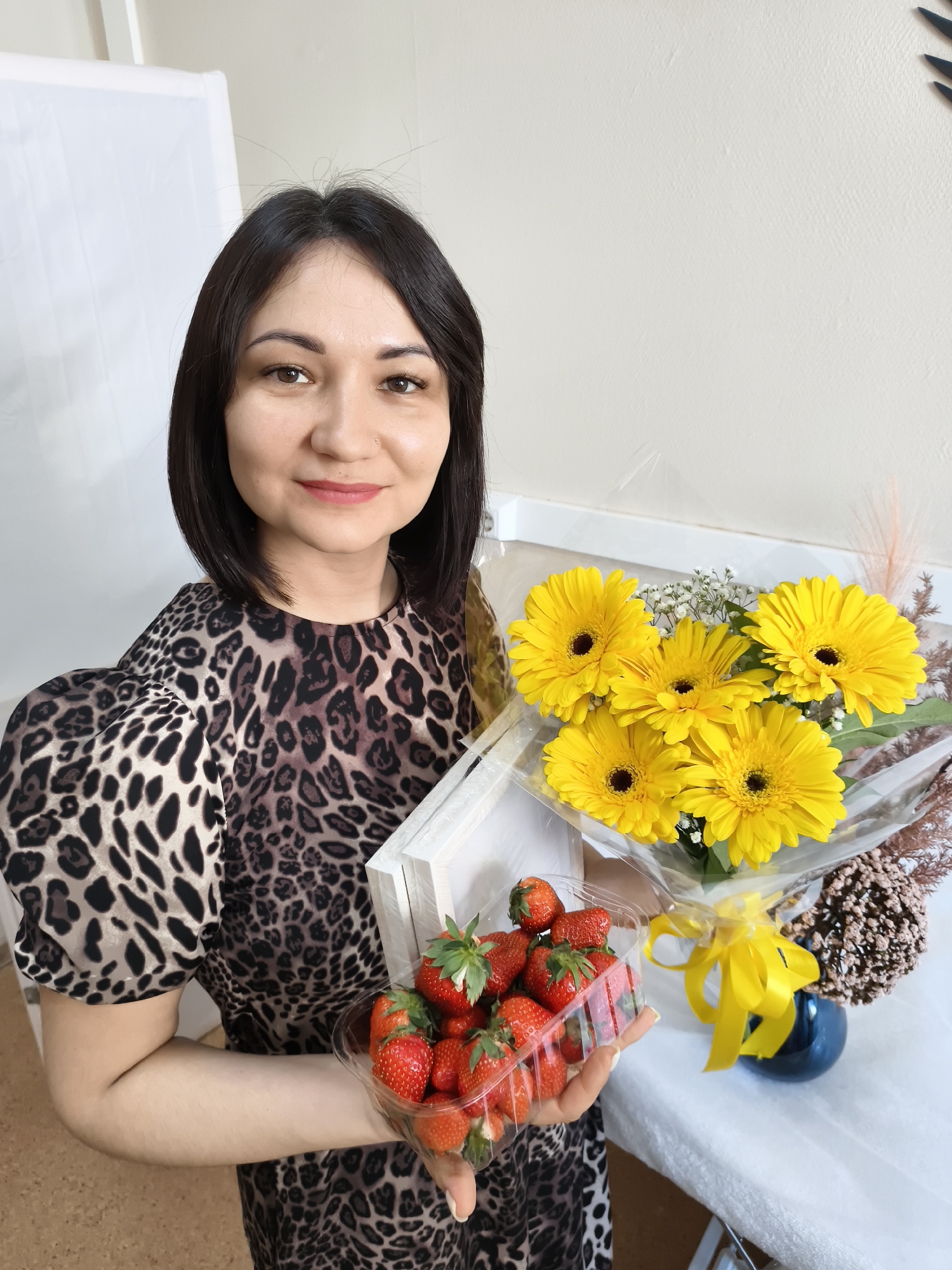 A woman with short, dark hair is holding a clear plastic container of strawberries and a bouquet of yellow gerbera daisies. She is wearing a brown and black leopard-print dress with puffy sleeves. The bouquet of flowers is wrapped in clear plastic and tied with a yellow ribbon. On the table to the right of the bouquet is a blue vase and a decorative pinecone-like object. The background consists of a white wall and a white folding screen. The location appears to be indoors.