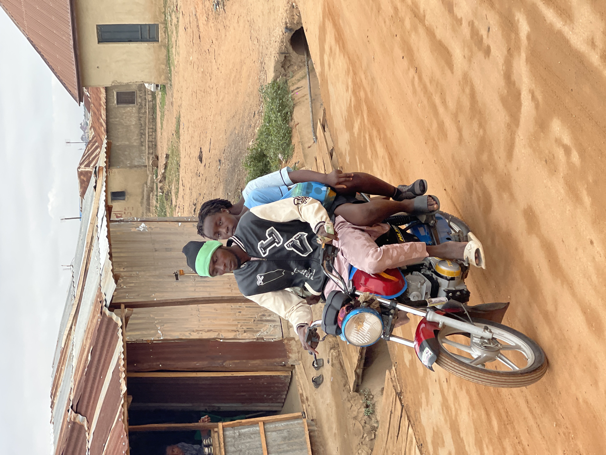 Two individuals are on a red motorcycle on a dirt road in Kuchingoro, Nigeria. The driver, wearing a black and white jacket and a green beanie, is looking directly at the camera. A second person is lying horizontally behind the driver. Buildings with corrugated metal roofs are visible in the background. The location appears to be a residential area with unpaved roads.