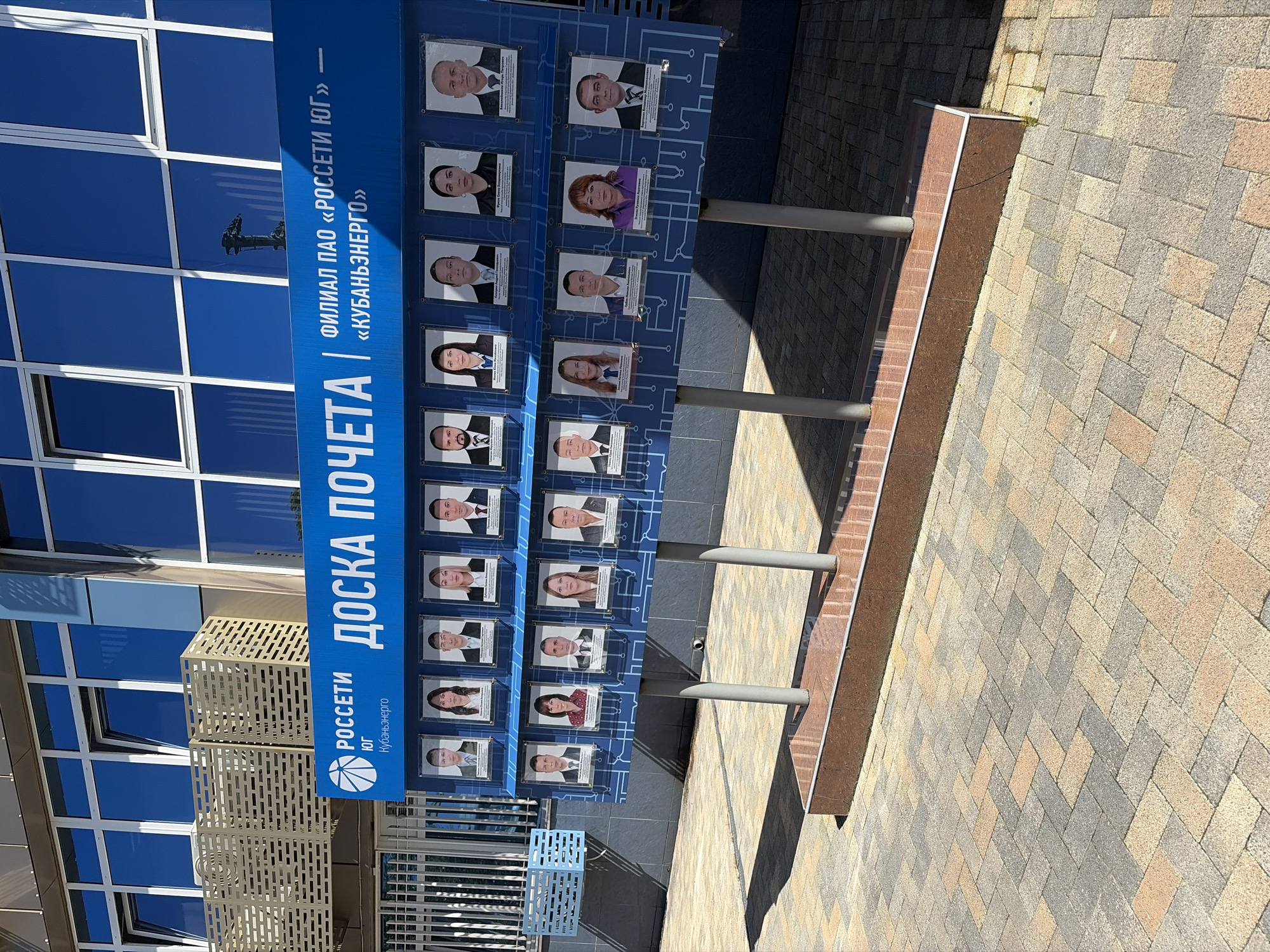 This image captures an outdoor "Board of Honor" (ДОСКА ПОЧЕТА) in Sochi, Russia, on a bright, sunny day. The board, which is a prominent blue structure, displays framed portrait photographs of approximately 20-24 individuals, likely employees of the organization. Each portrait is accompanied by a small label, presumably detailing the person's name and achievements. The blue background of the board features a subtle circuit board-like design.
The visible text on the board identifies the organization as "ФИЛИАЛ ПАО «РОССЕТИ ЮГ»" (Branch of PJSC "Rosseti Yug") and "«КУБАНЬЭНЕРГО»" ("Kubanenergo"), with a distinctive logo for "РОССЕТИ ЮГ Кубаньэнерго" prominently displayed.
Behind the board, part of a modern building with large, blue-tinted, reflective glass windows is visible, reflecting the clear sky. The board is supported by grey metal poles, which cast distinct shadows onto the ground, indicating direct sunlight. The ground in front of and around the board is paved with multi-colored interlocking pavers in shades of brown, grey, and beige, arranged in a herringbone pattern. A raised border made of dark brown material runs along the edge of the paved area. To the left, a light-colored, slatted metal structure, possibly a bench or decorative element, is partially visible. The scene suggests a professional or corporate setting outdoors.