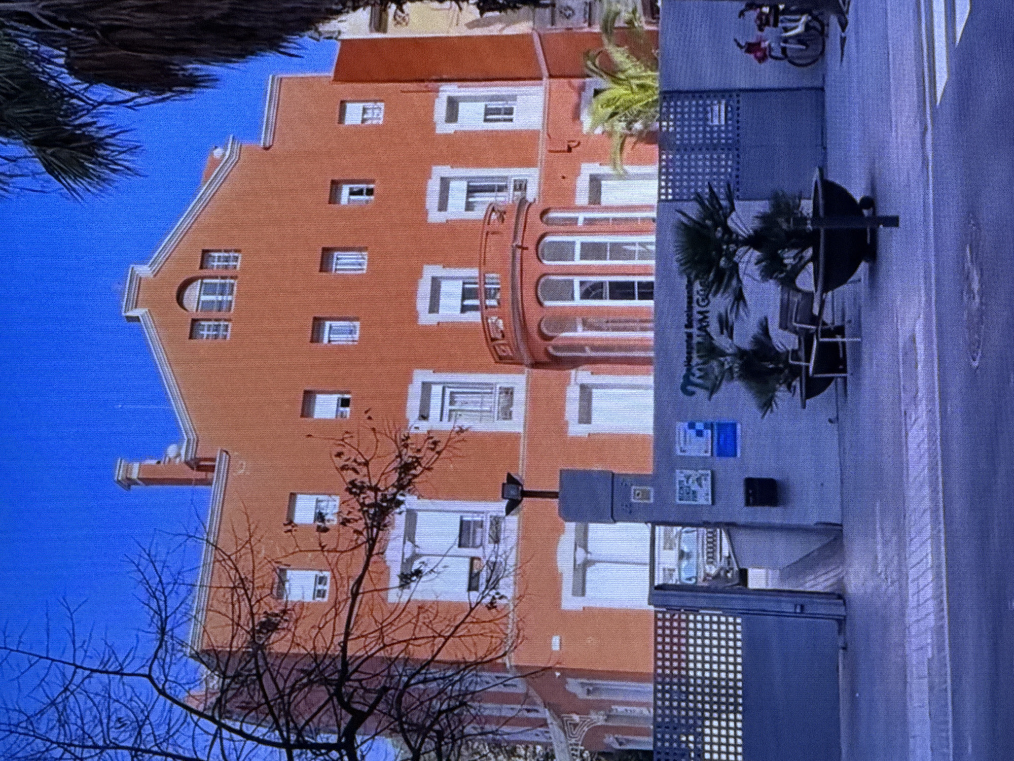 This is a vertical, close-up, eye-level shot of an orange building in Dubai. The building has numerous windows, some with white frames and others with dark frames. There's a curved balcony with windows and railings. A palm tree is visible on the right side of the building, and a dark-colored object, possibly a planter, is in front of it. Below the balcony, there's a fence with a repeating square pattern. The sky is bright blue, suggesting daytime. Bare branches of a tree are in the foreground on the left, partially obscuring the building. To the right, a section of a grey structure with square patterns and some signage is visible. The signage appears to have text in a light color, but it is difficult to read clearly due to the angle and resolution. The overall impression is a sunny day in an urban environment.
