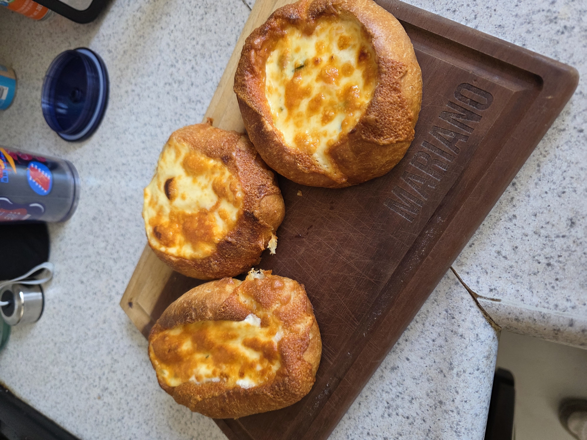 Three freshly baked items, resembling bread bowls or rolls filled with a golden-brown, bubbly cheese mixture, are prominently displayed on a dark wooden cutting board. The largest item, positioned at the top right, is a round bread bowl with a rich, melted cheese filling, showing some green specks, possibly herbs. Below it, two other similarly filled and baked items, slightly elongated, rest on the board. All three have a crusty, golden-brown bread exterior, indicating they are likely straight from the oven.
The wooden cutting board features a carved inscription, "MARIANO," in capital letters along its right side. It has a groove along one edge, typical for collecting juices. The setting appears to be a kitchen or dining area, with a speckled, light-colored countertop serving as the background surface. In the upper left background, part of a blue plastic lid and a colorful tumbler or bottle are visible. A metallic object, possibly a kitchen utensil, is partially seen on the far left. The lighting suggests daytime, illuminating the food and surfaces clearly. No human presence or activity is depicted, but the arrangement implies a meal is prepared or about to be served. The overall impression is one of warm, homemade comfort food.