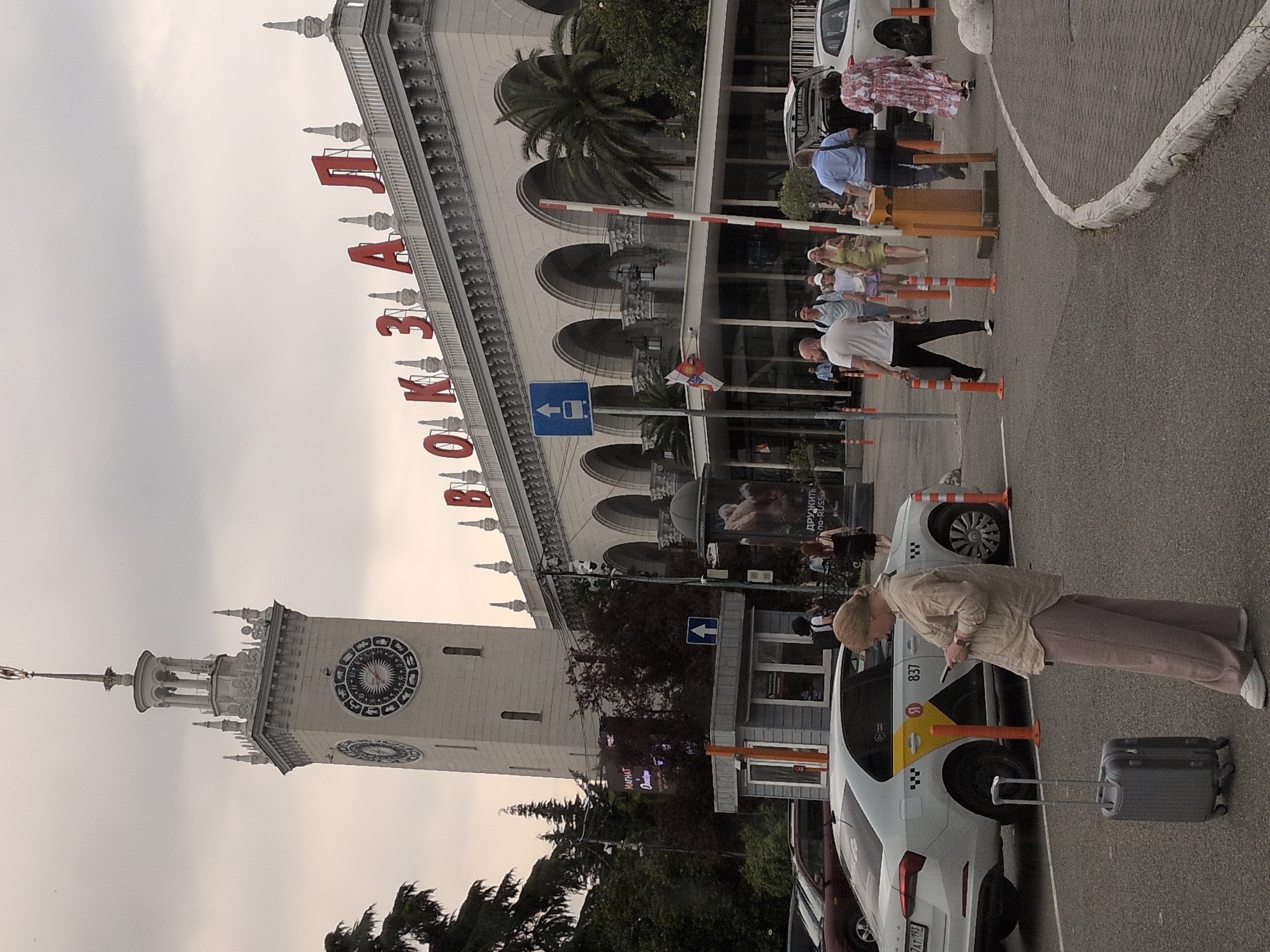 The image shows a view of the Sochi railway station in Russia, a large, ornate building with a prominent clock tower. The sky is overcast, and the scene appears to be outdoors in what looks like a transportation hub. In the foreground, a woman with blonde hair is standing, looking at her phone, with a gray rolling suitcase next to her. She is wearing a light-colored, loose-fitting top and loose pants. A white taxi with yellow accents and the number "837" is parked nearby. Several other people are visible in the background, walking or standing, including a group of people near a yellow barrier gate. The architecture of the station is grand, with arched windows and decorative elements. The word "ВОКЗАЛ" (Vokzal), meaning "station," is prominently displayed in large red letters across the top of the building. Palm trees are visible near the station, suggesting a warm climate. The overall atmosphere seems busy but not chaotic.