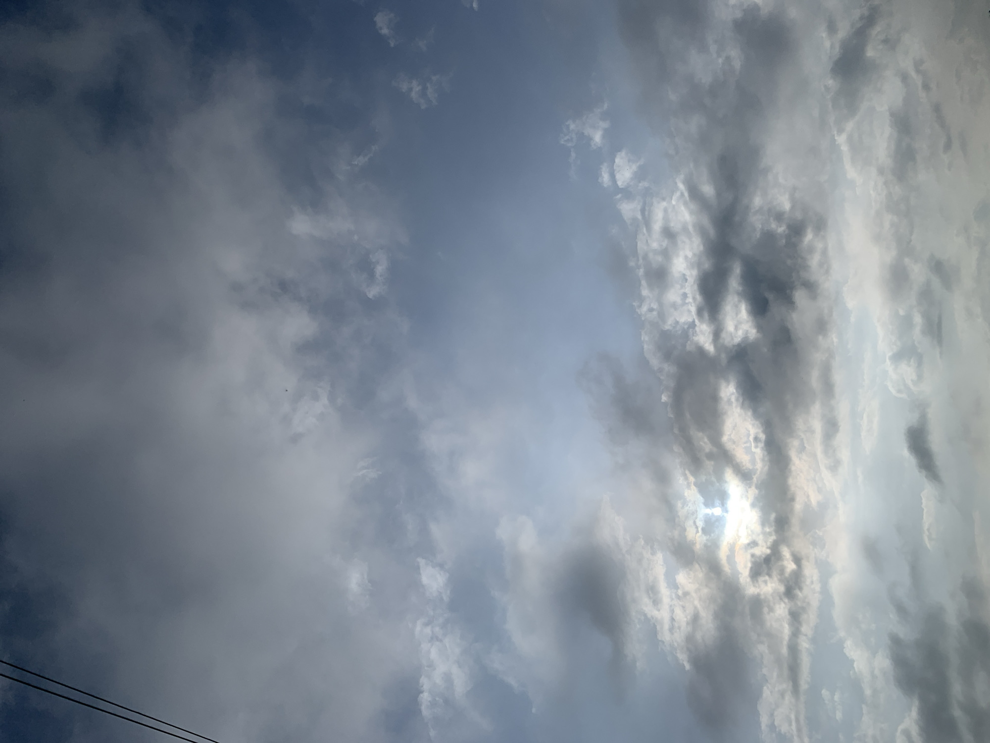 The image captures a cloudy sky with the sun partially obscured by thick, gray clouds. The sky transitions from a pale blue at the top to a brighter, more illuminated area near the horizon where the sun's rays are breaking through. Wisps of white clouds are scattered throughout the frame, some appearing thin and translucent, while others are dense and heavy with moisture. Two dark, thin lines, likely power lines, are visible in the upper left corner of the image, bisecting the sky. The overall scene suggests an overcast day with intermittent sunlight. The location is Kuru Karama, Nigeria.