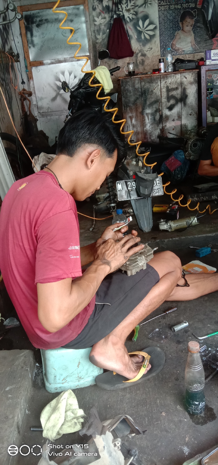A young man, likely a mechanic, is engrossed in repairing a motorcycle engine part inside what appears to be a cluttered garage or workshop in Pesawahan, Indonesia. He is positioned on a light blue plastic stool, wearing a red t-shirt and black shorts, with one foot bare and the other wearing a flip-flop. His hands are visibly stained with grease as he meticulously works on the metal engine component.

Behind him, a motorcycle is partially visible, featuring a license plate that reads "N 29 JE" with additional numbers below it, likely "27" or "99 JE 27" based on context. Above, a coiled yellow air hose hangs. The workshop walls are adorned with stenciled leaf patterns and show signs of wear. A wooden counter in the background holds various items, including a mug, bottles, and boxes, along with a red drawstring bag hanging from the wall. A small picture of a baby and a glass pane with faint writing, possibly "Mobile heges," are also visible. An angle grinder tool rests on the floor nearby, among other scattered tools like a screwdriver and wrenches. A clear bottle containing a green liquid is also on the ground.

The scene depicts a typical working environment for vehicle repair, with the man deeply focused on his task, suggesting an active repair or maintenance session during the daytime. Visible text includes a phone number "CALL 08121946, 4038" on a sign and the brand "AIMNO" on the man's shirt. The watermark "Shot on Y15 Vivo AI camera" is in the bottom left corner.