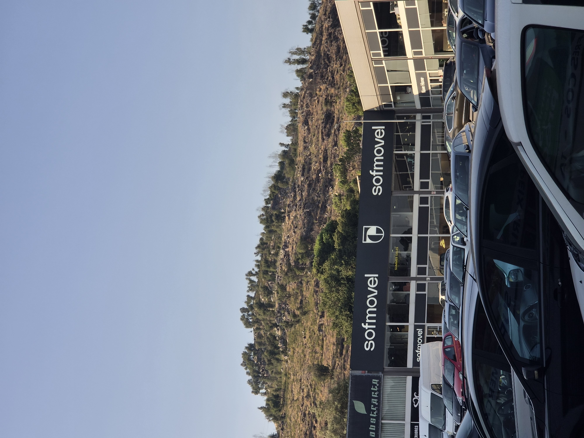 The image depicts a daytime scene outside a commercial building in Braga, Portugal. The building features prominent signage for "Sofmovel" and "Abstracta Interiors". Several cars are parked in a lot in front of the building. In the background, a hillside covered with vegetation and rocks is visible under a clear blue sky. The architectural style of the building suggests a retail or office space. No people are clearly visible in the image, and no specific actions or interactions are depicted. The overall environment appears to be a commercial area on the outskirts of the city.