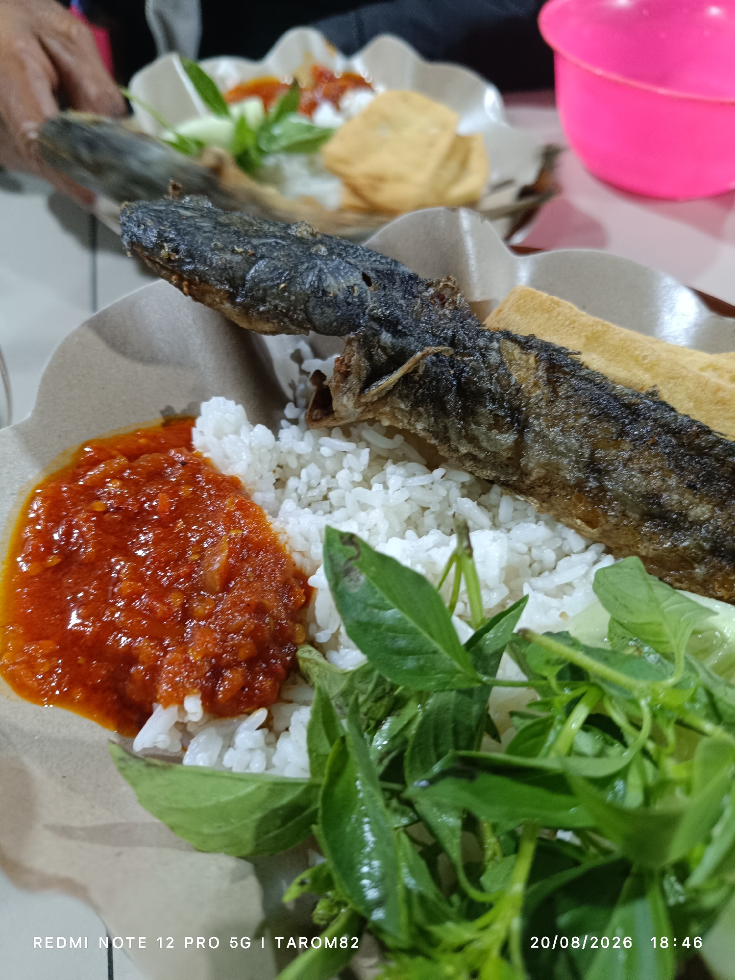 In Bringinbendo, Indonesia, a close-up, eye-level shot captures a plate of Indonesian cuisine. The dish features a whole fried fish, white rice, a dollop of vibrant red sambal (chili paste), and a side of green vegetables, possibly basil. Two pieces of fried tofu are also visible on the plate. The meal is served on a brown disposable plate with a scalloped edge. In the background, a person's hand is partially visible, reaching for something. A bright pink bowl is present in the upper right corner of the frame. The bottom of the image displays a timestamp, "20/08/2026 18:46," and text, "REDMI NOTE 12 PRO 5G | TAROM82." The lighting suggests an indoor setting, likely a restaurant.